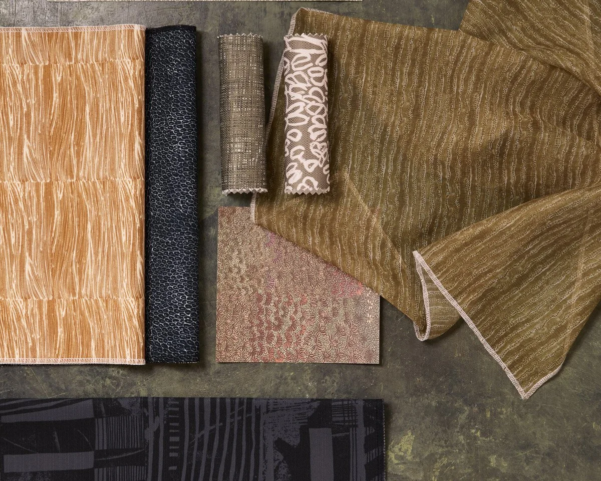 Various patterned fabrics laid out and folded on a flat grey surface, in earthy pink, green and gold colours.