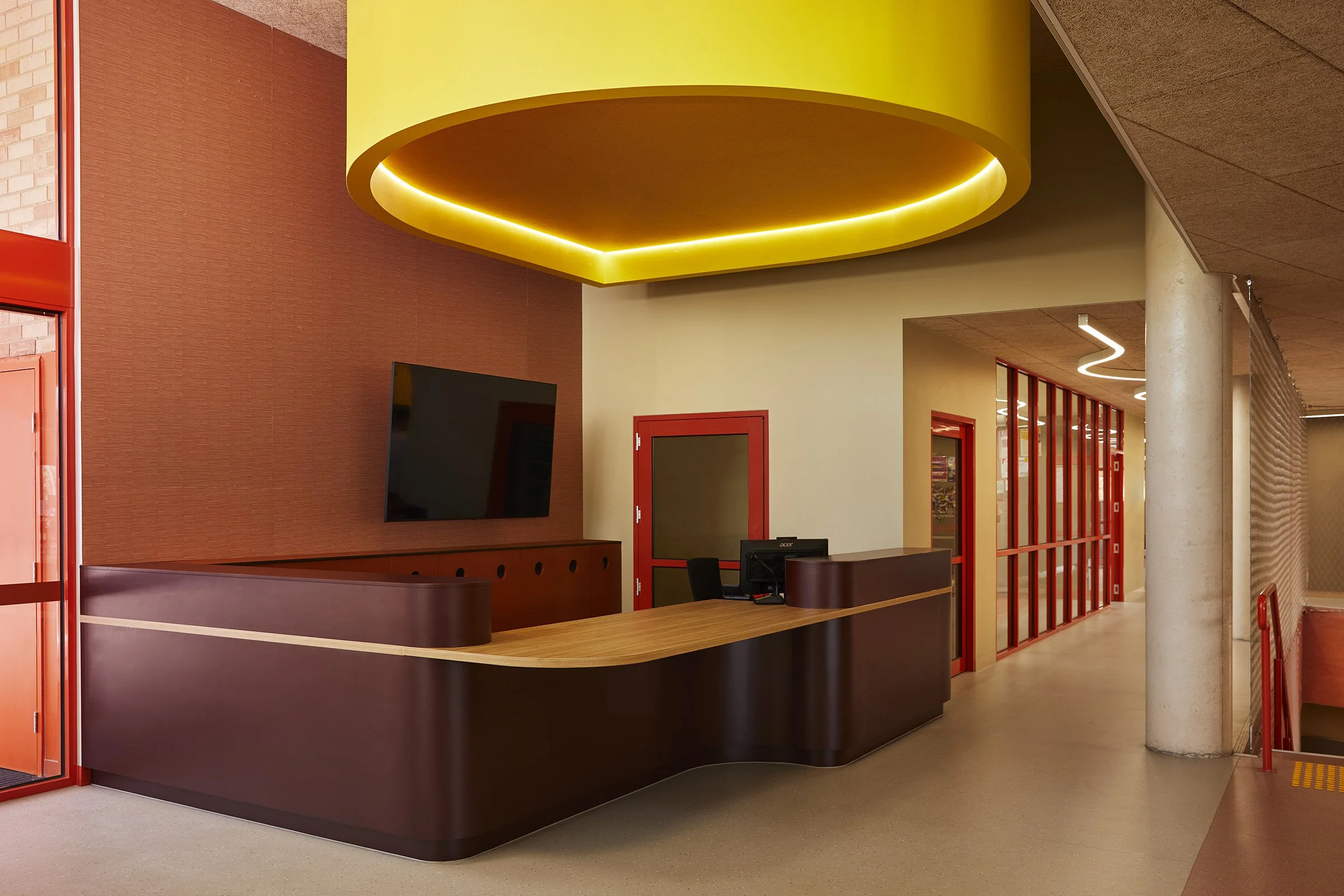 A bold reception space for Auburn High School, featuring printed acoustic panelling in muted orange tones next to red doorways, a timber and deep purple coloured wrap around desk. A large yellow bulkhead sits above the desk and emits subtle light.