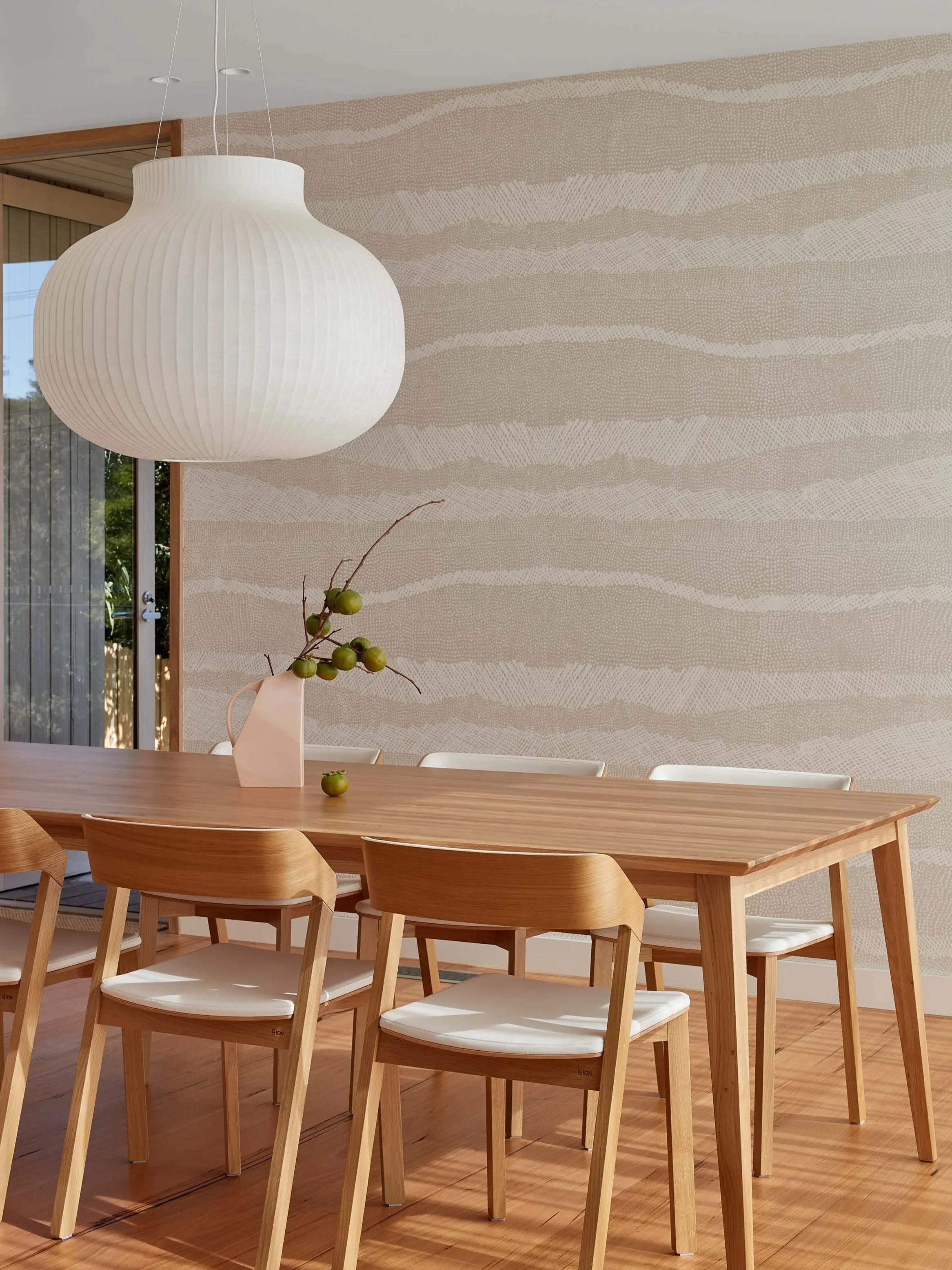 The dining space of Blairgowrie Beach Club features a stone coloured patterned wallpaper. A large paper pendant light hangs over the timber dining table with a pale pink vase. The table, chairs and timber floors are drenched in sunlight.