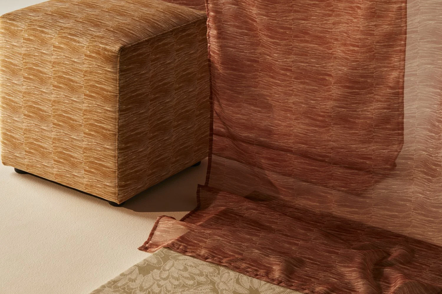 A piece of patterned fabric with a floral design, next to a sheer, brownish-red curtain, with a piece of fabric matching the curtain's color on the chair.