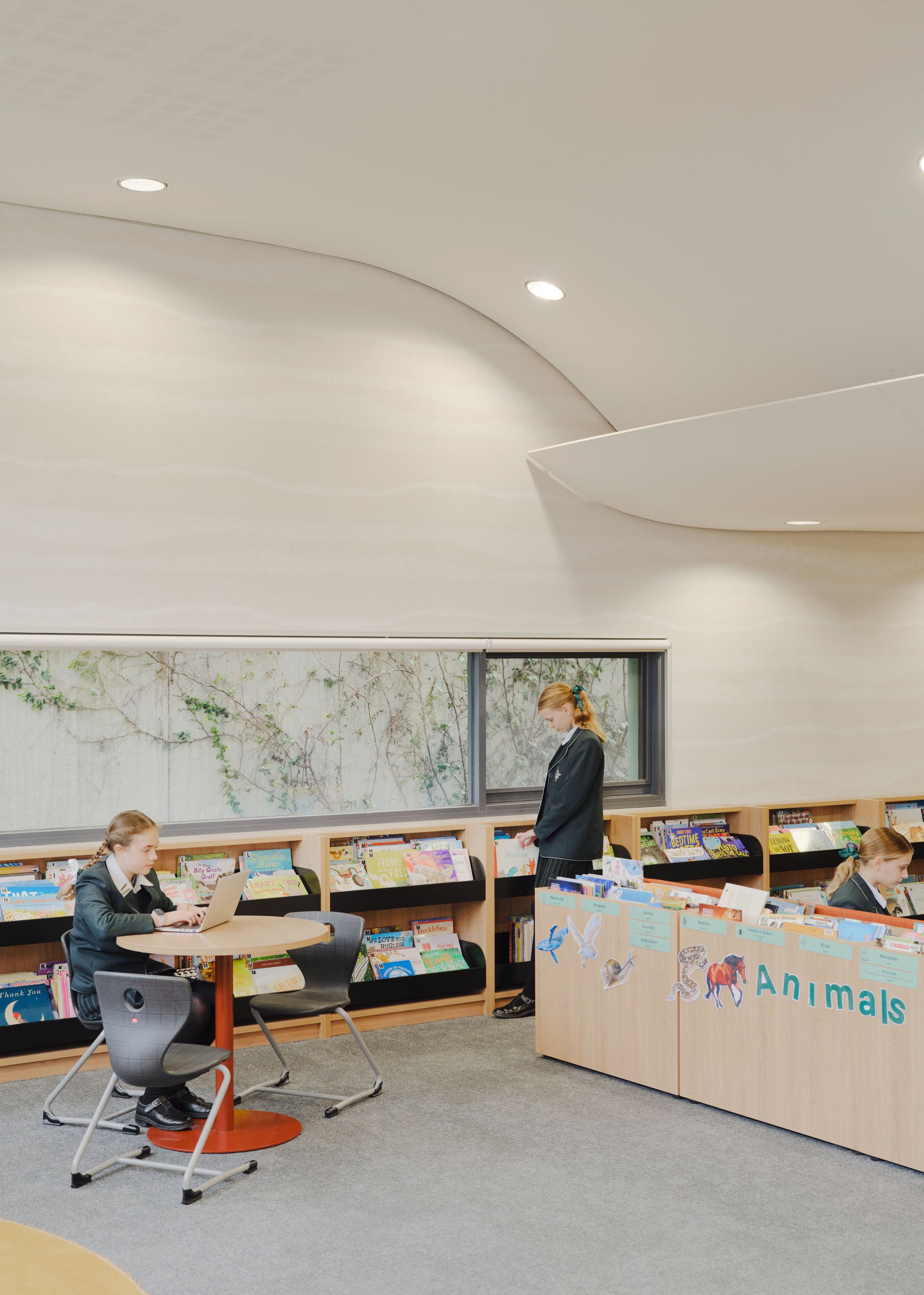 Students in school uniforms in a library reading books and using a tablet. The library has a display with animals and the word 'Animals' visible.