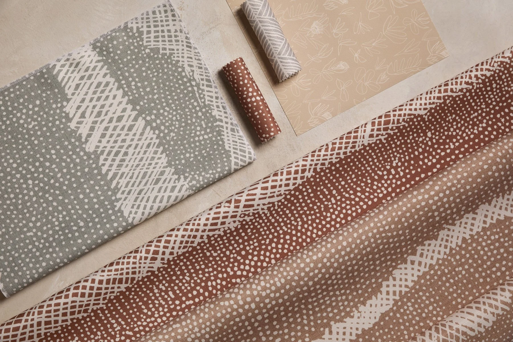 Various patterned fabrics and wallpapers laid out and folded on a flat surface, in earthy pink, green and sandy colours.