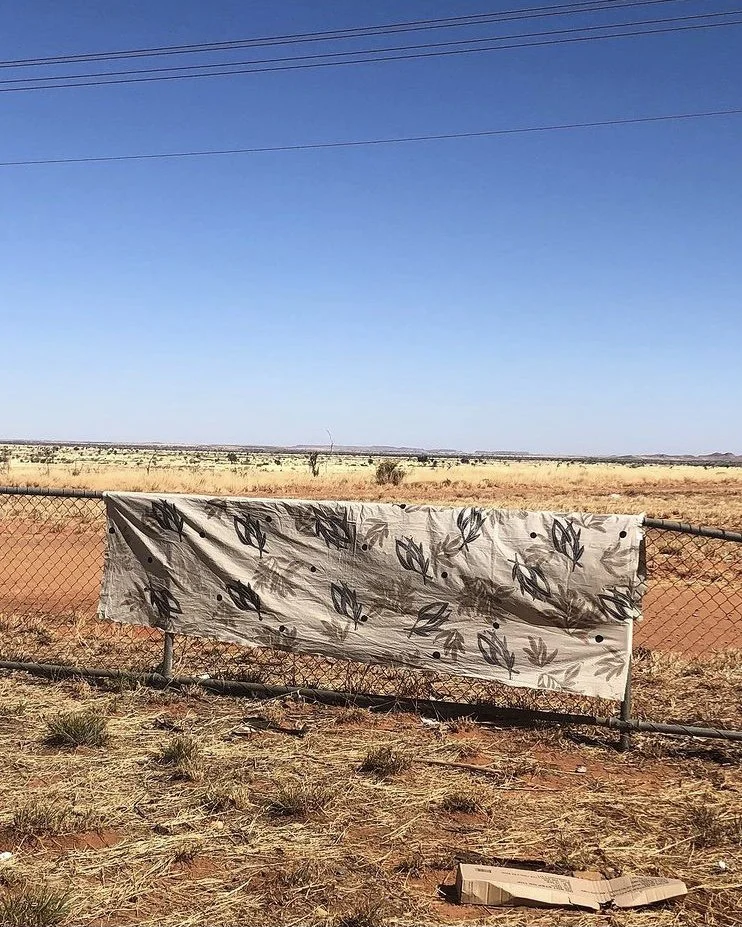 Australian landscape with wire fence with printed fabric hanging over it, bright blue sky with red soil and grass behind.