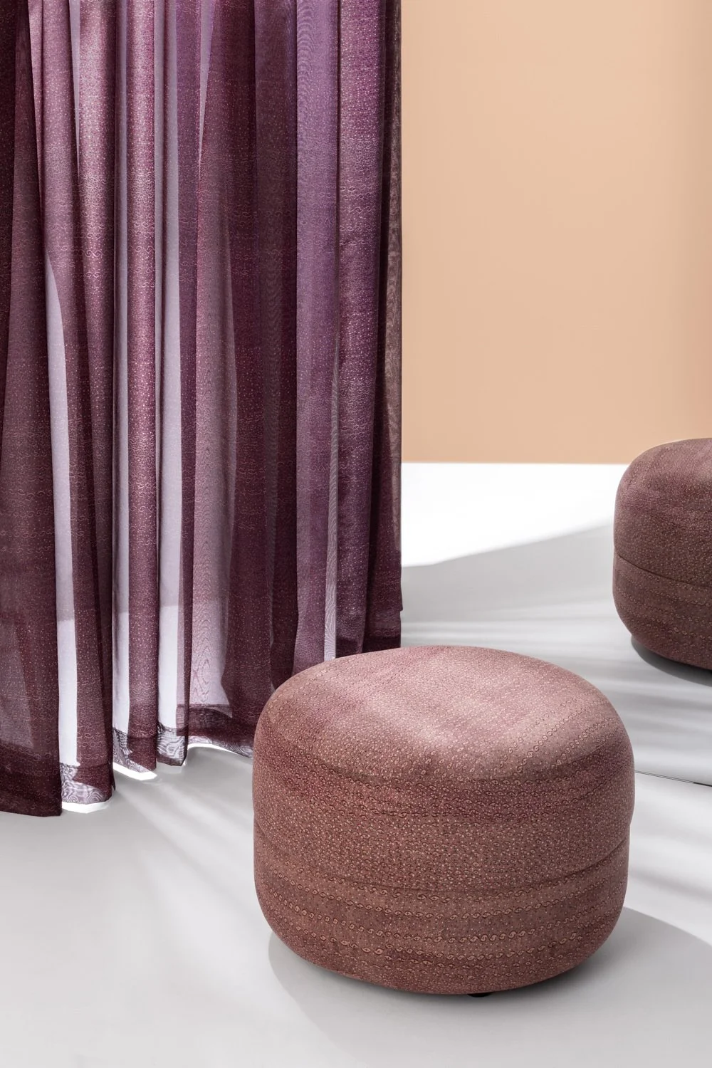 Interior with round ottoman and sheer drapery curtain with patterned fabrics featuring First Nations designs with colours of pink and plum, with ottoman reflected in mirror and a pale peach coloured wall