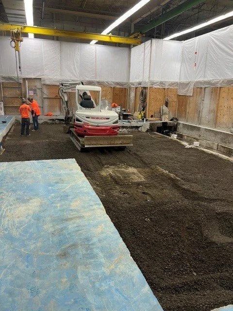 Construction site with a compacting machine, workers, and a crane in an unfinished indoor structure.