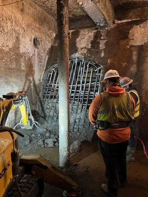 Two construction workers in safety gear inspecting a damaged concrete structure with exposed rebar in an underground space.