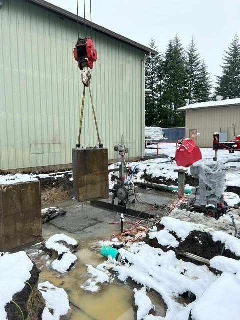 Construction site with a crane hoisting a large metal block over a hole in the ground, snow on the ground, and construction equipment nearby.