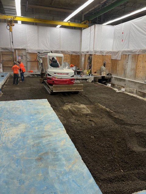 Construction site with a small excavator smoothing dark dirt, two workers in orange shirts, and construction materials around.