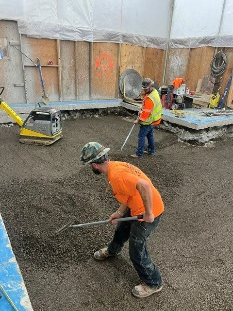 Two construction workers leveling gravel on a construction site, with tools and equipment around them.