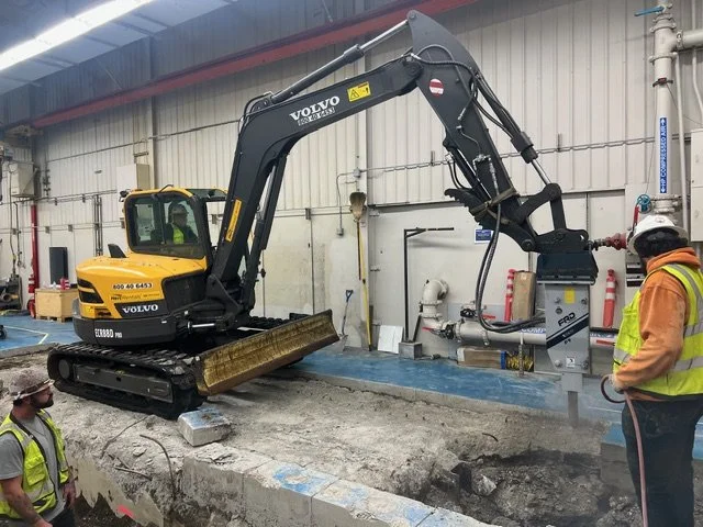 A yellow Volvo mini excavator working inside an industrial facility, with two workers in safety gear nearby.