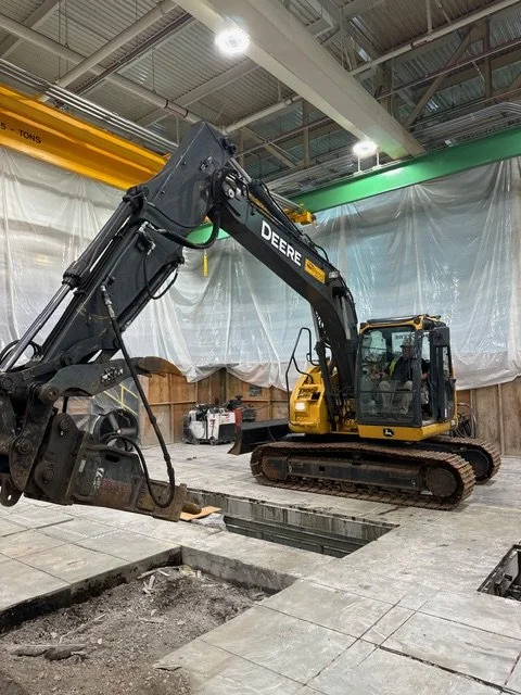 A John Deere excavator inside a construction site is lifting a concrete slab. The site has an overhead crane and partially finished flooring.