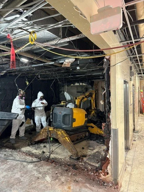 Construction workers in protective suits inspecting a damaged interior with exposed ceiling and a small excavator. Part of the wall is demolished.