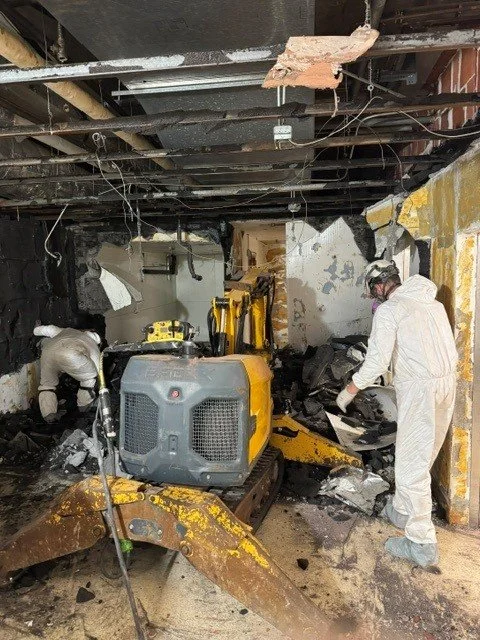 Two people wearing protective gear work inside a burned and damaged building with exposed ceiling beams and charred walls, using a small yellow excavator to clear debris.