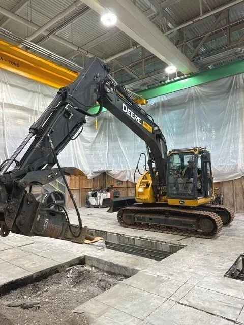 A John Deere excavator pinpointing a section inside a warehouse or factory with concrete flooring and overhead lighting.