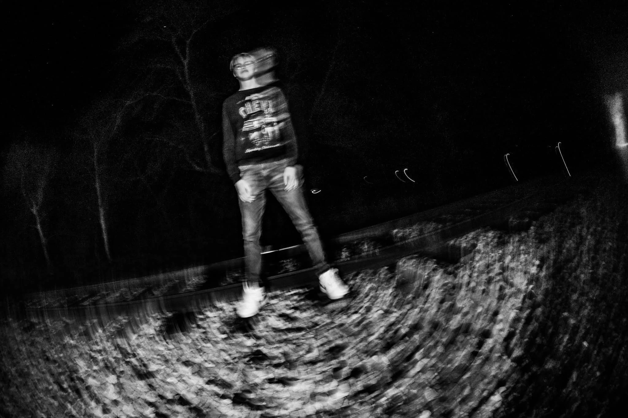 A blurry black-and-white photo of a person standing on train tracks at night, wearing glasses, a sweatshirt, jeans, and boots, with trees and streetlights in the background.