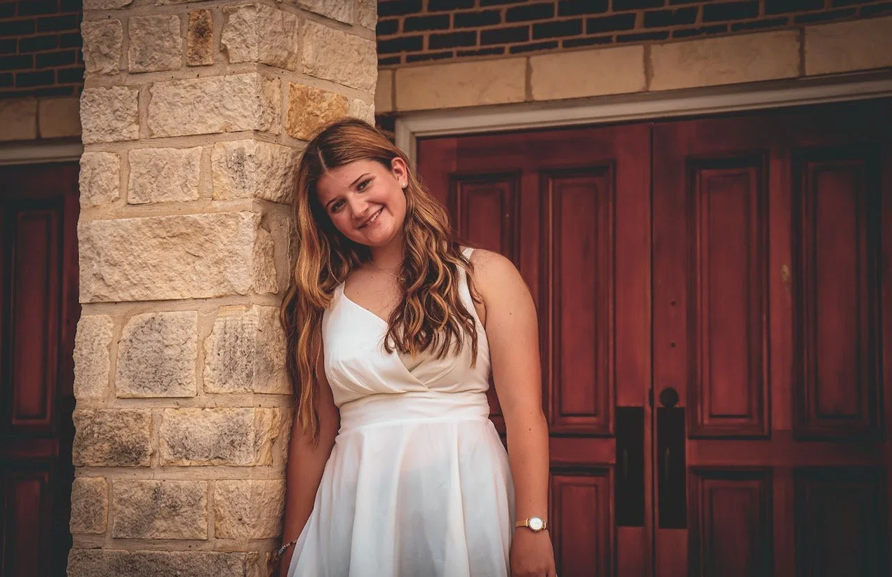 A young woman with long, wavy brown hair smiling and leaning against a stone pillar outside a building with dark wood doors.