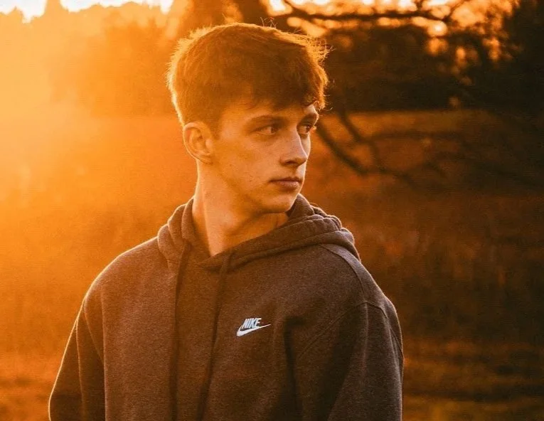 A young man wearing a dark Nike hoodie outdoors during sunset, with trees and orange sky in the background.