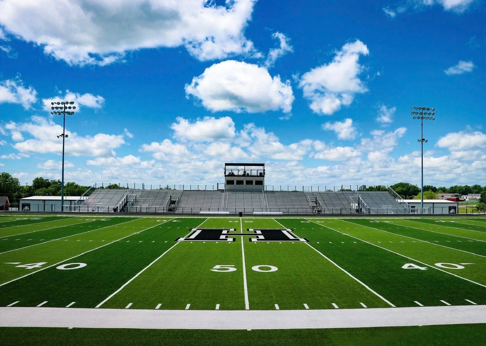 Empty football field with green turf, white yard lines, and the letters T.G.I. on the 50-yard line. Bleachers, light poles, and a sky with scattered clouds are visible in the background.