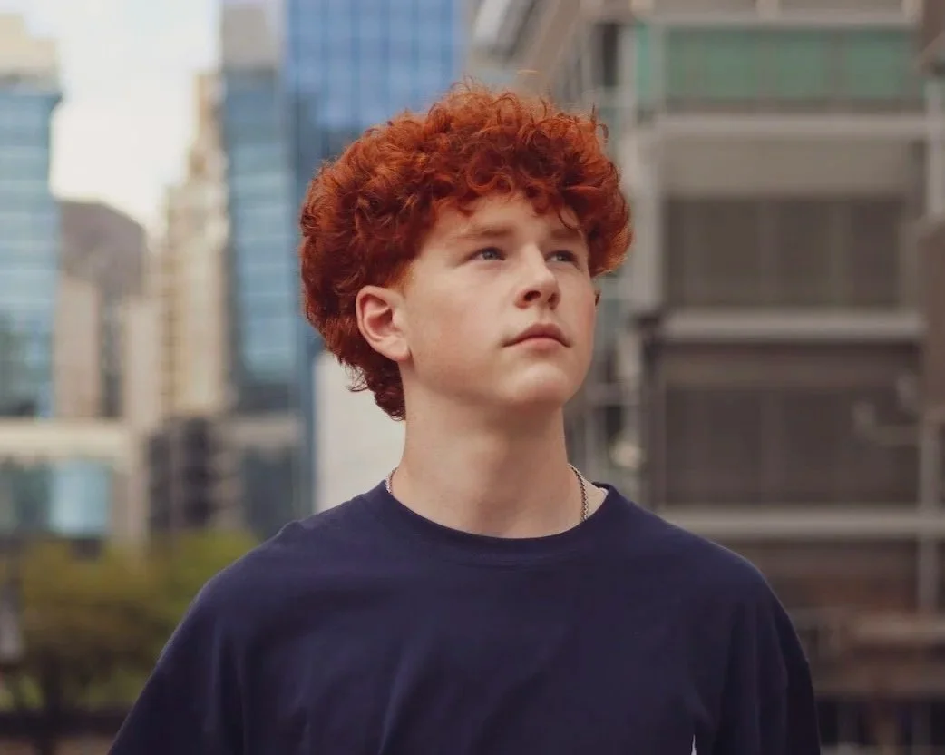 A young person with curly red hair wearing a dark blue shirt stands outdoors in an urban area with tall buildings in the background.