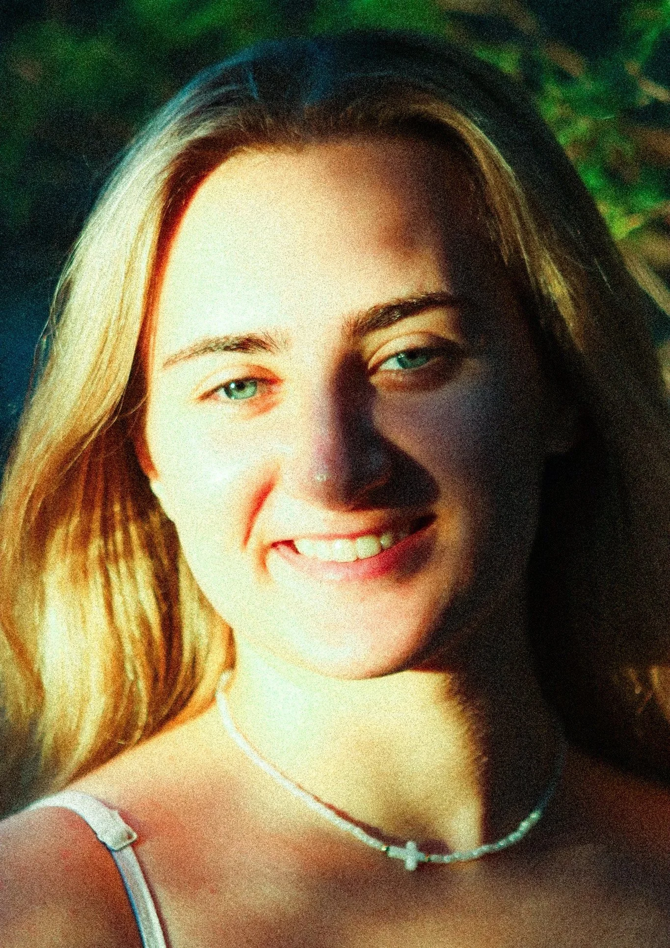 A young woman with blonde hair and blue eyes smiling outdoors with greenery in the background.