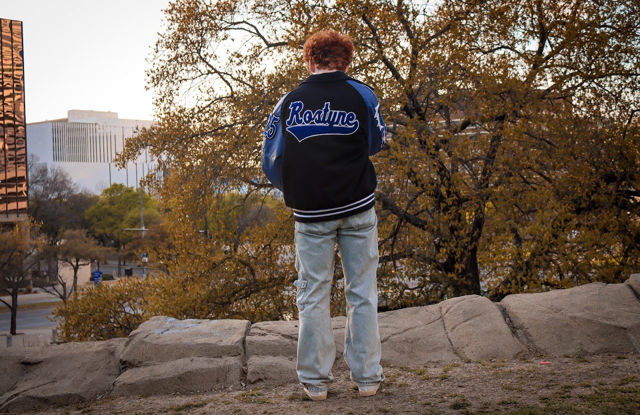 Person with red hair wearing a black and blue varsity jacket and light jeans, standing on rocks and looking at trees with autumn foliage.
