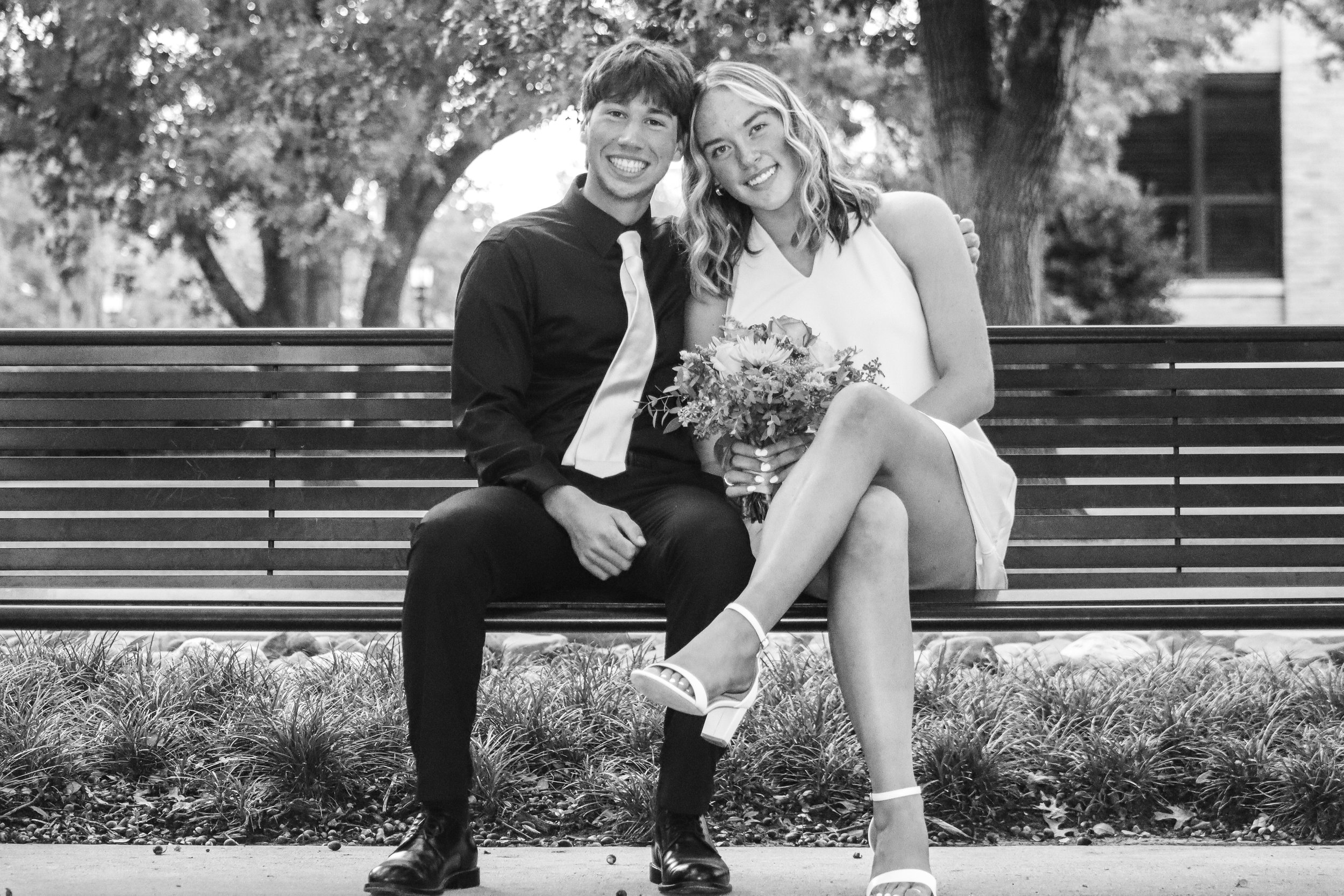 A young couple sitting on a park bench, smiling. The woman is holding a bouquet of flowers, wearing a white dress and white high heels. The man is in a dark shirt and tie. They appear happy and relaxed, with trees in the background.