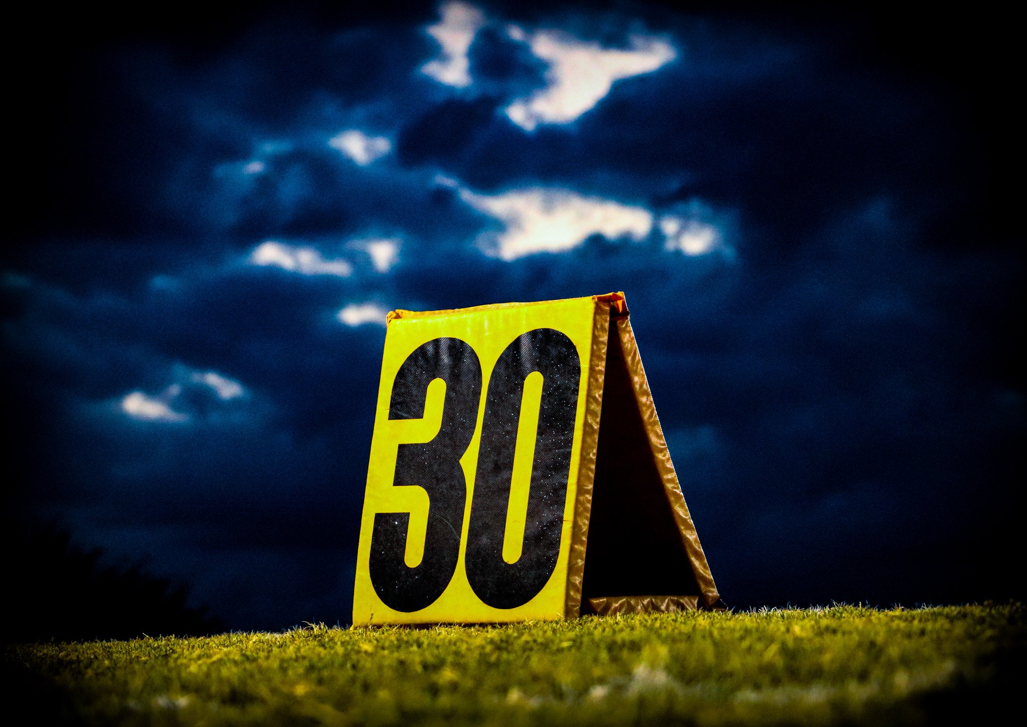 A bright yellow sign with black numbers reading 30, standing on grass against a dark, cloudy sky at dusk.
