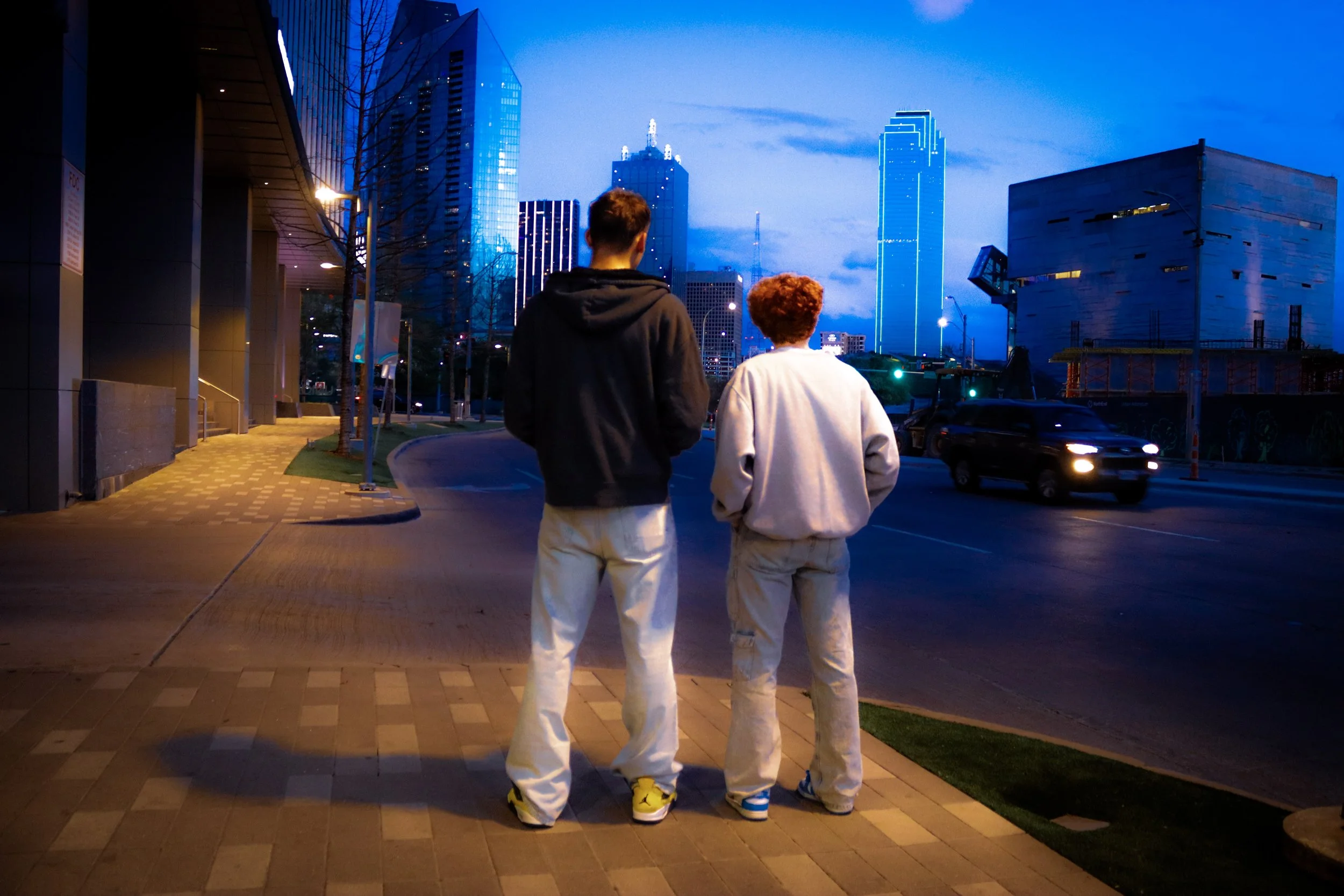 Two young people stand on a city sidewalk at dusk, facing away from the camera. They are near a street with moving cars, with tall illuminated buildings and a partly cloudy sky in the background.