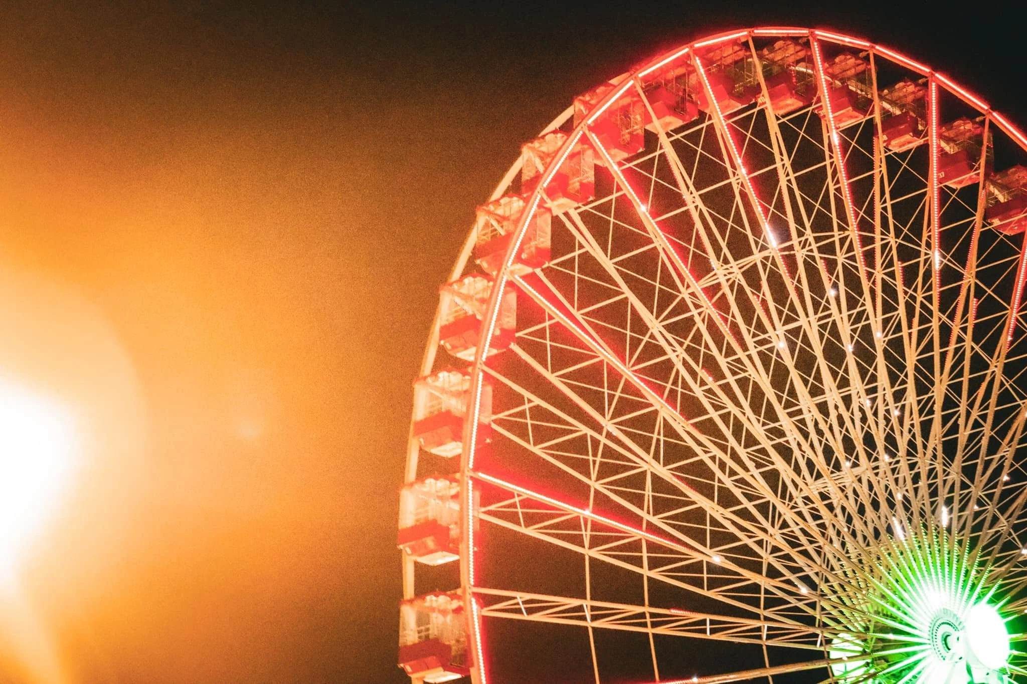 Night view of a brightly illuminated Ferris wheel with red lights, showing part of the wheel's structure and green lights at the center, with a bright, overexposed light source on the left side.