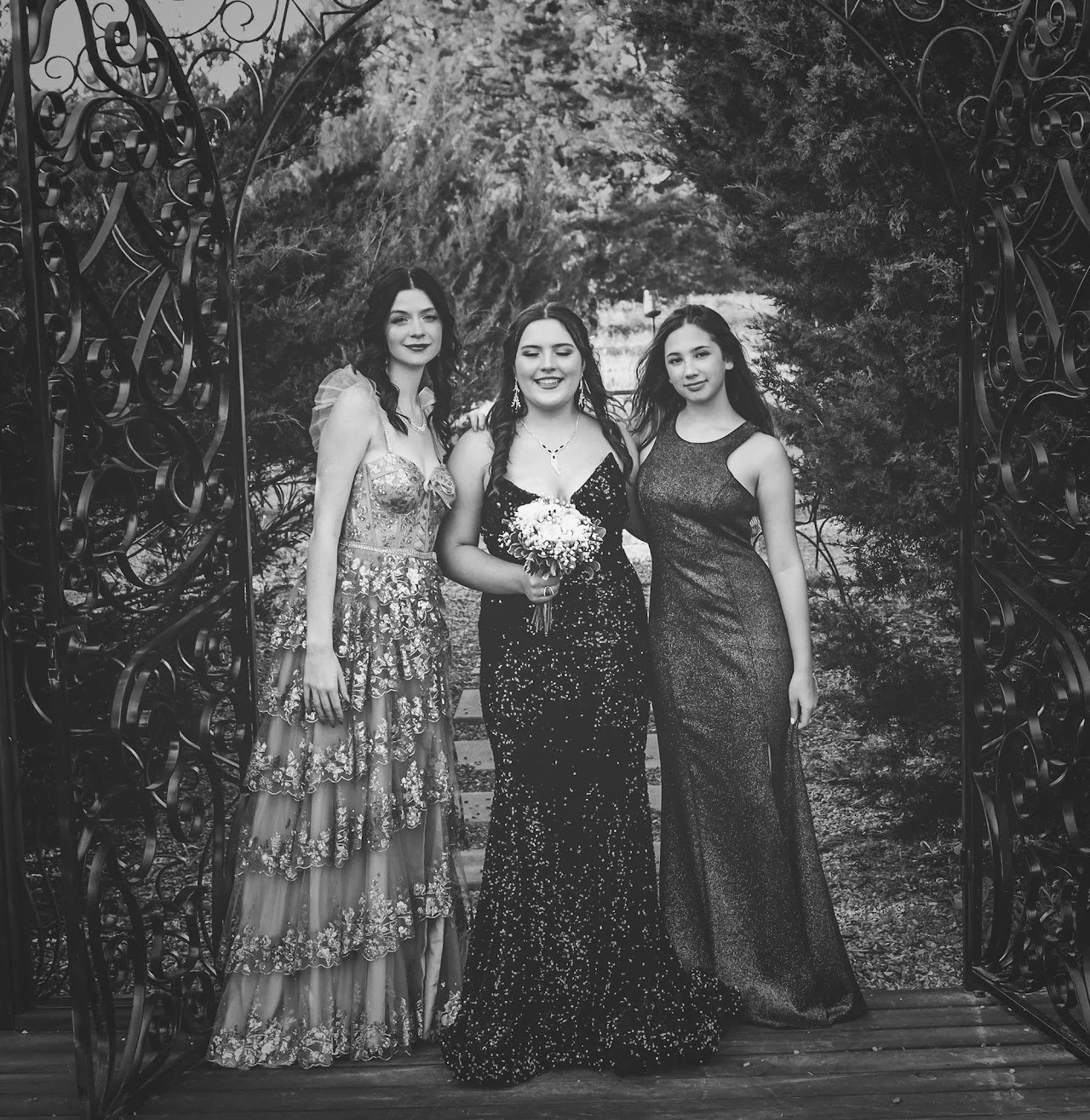 Three women in formal dresses standing together outdoors, one woman in the center holds a bouquet, posing beneath a decorative archway with trees in the background.