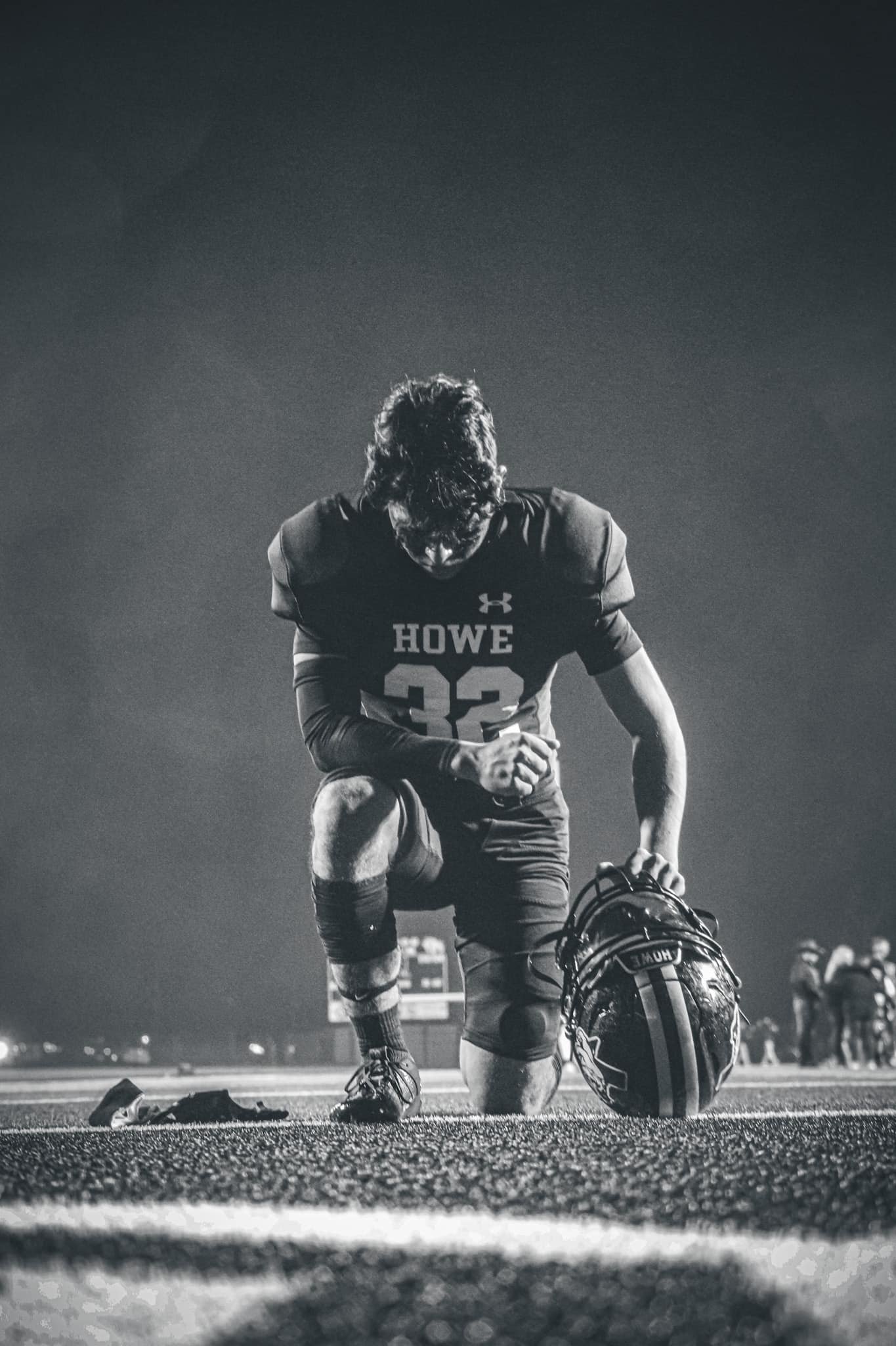 A football player kneeling on the field holding his helmet during night game, dressed in uniform with the word 'HOWE' and the number 32, at a sports stadium.