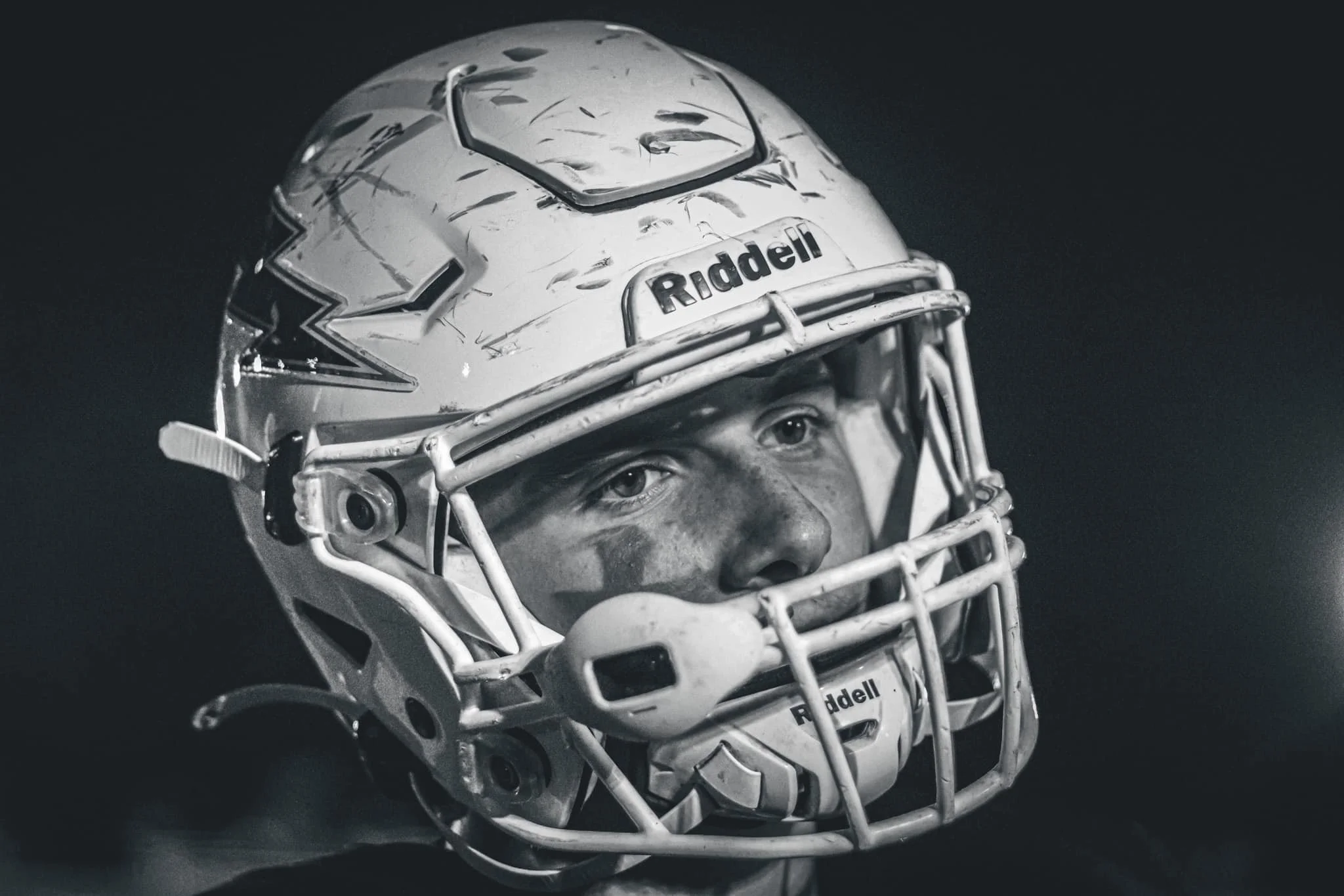 A close-up black and white photograph of a football player wearing a helmet with a faceguard, with the brand name 'Riddell' visible on the front.