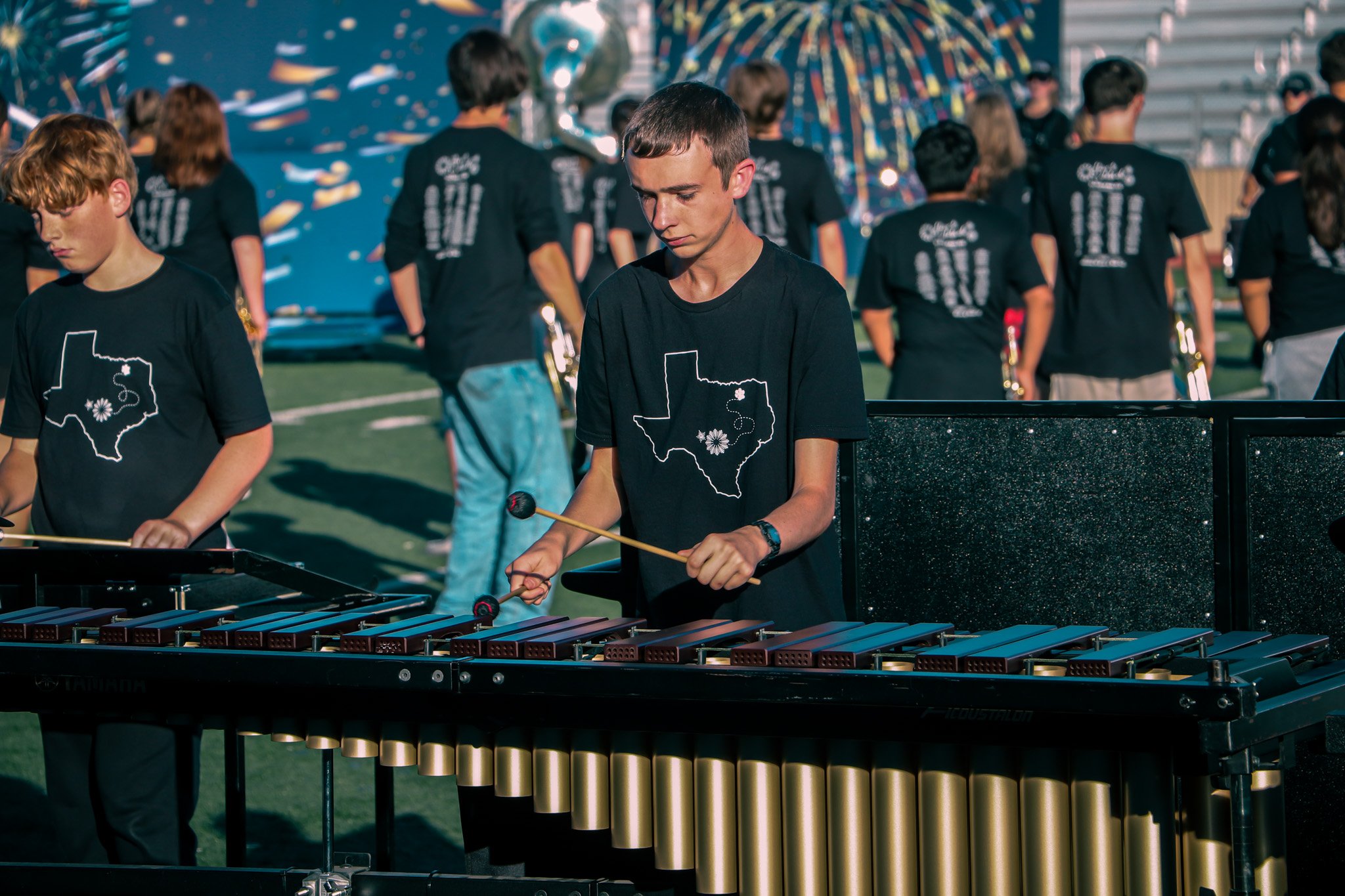 A young male musician playing a marimba during a band practice on a football field, with other band members in the background.