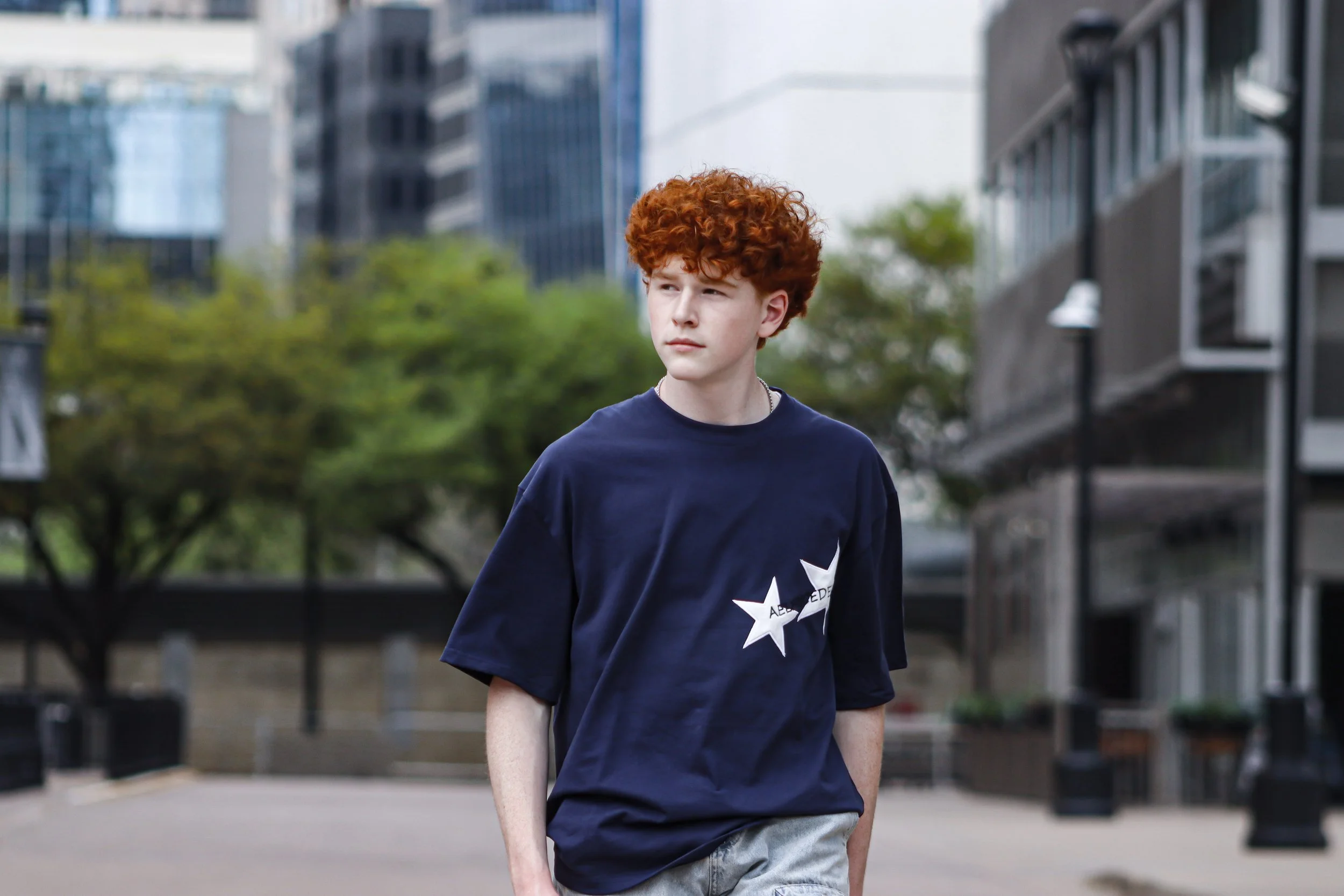 A young person with curly red hair walking outdoors in an urban area with tall buildings and trees in the background.
