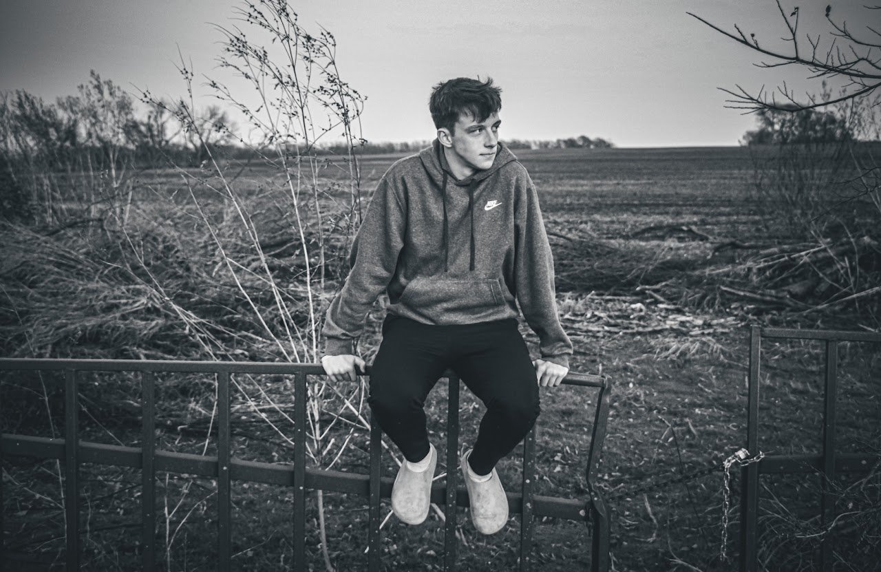 A young man sitting on a metal gate in a rural field, looking off into the distance, with bare trees and an overcast sky around him in black and white.