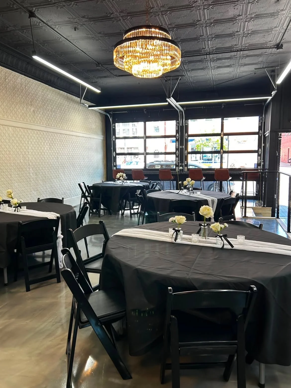 A decorated event space with round tables covered in black tablecloths, each with a small floral centerpieces and white runners, set up for a celebration or dinner, featuring large windows, a dark ceiling with a chandelier, and modern lighting.