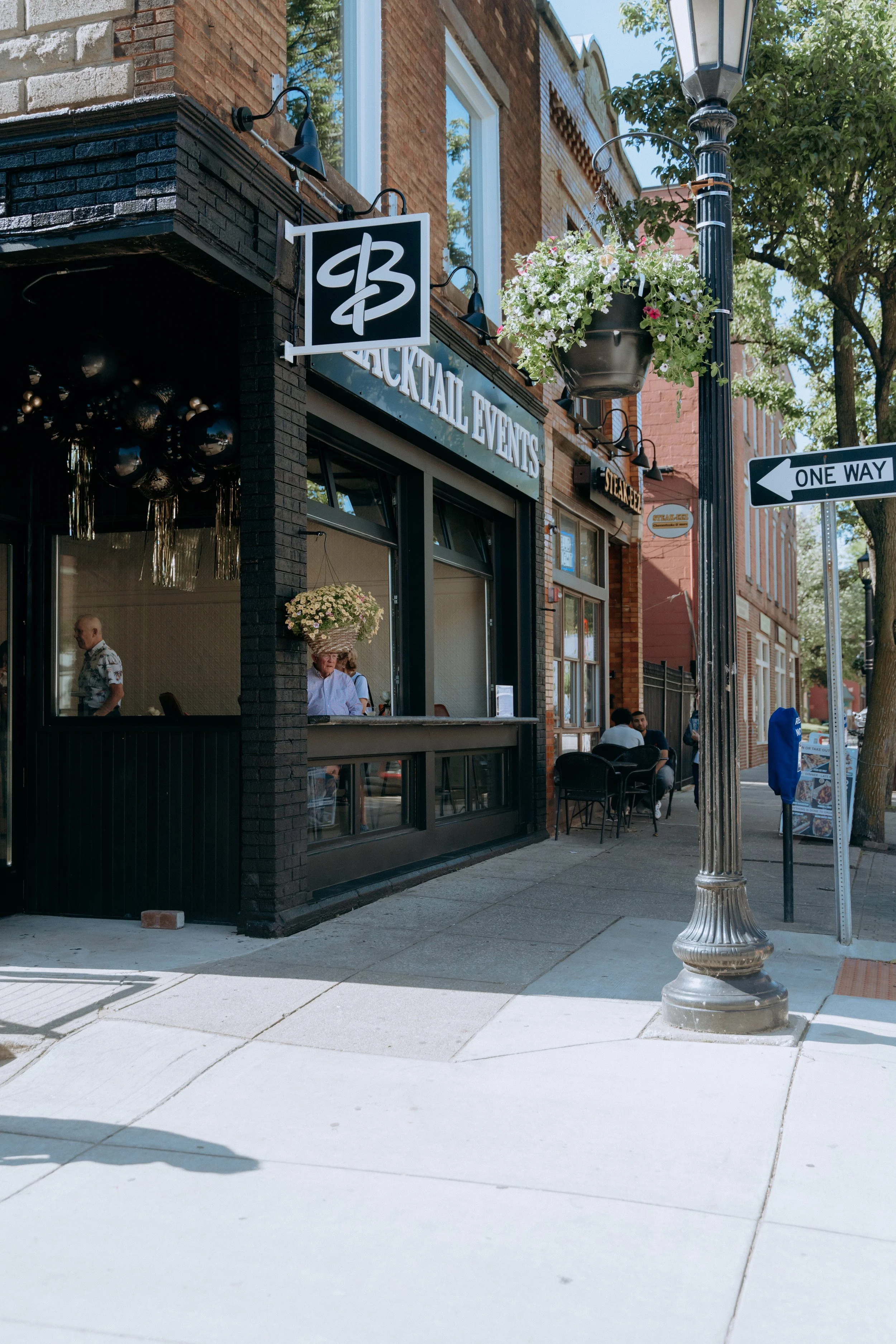 Street view of Blacktail Events with a black brick facade, hanging flower basket, street lamp, one-way sign, and people sitting outside and inside.