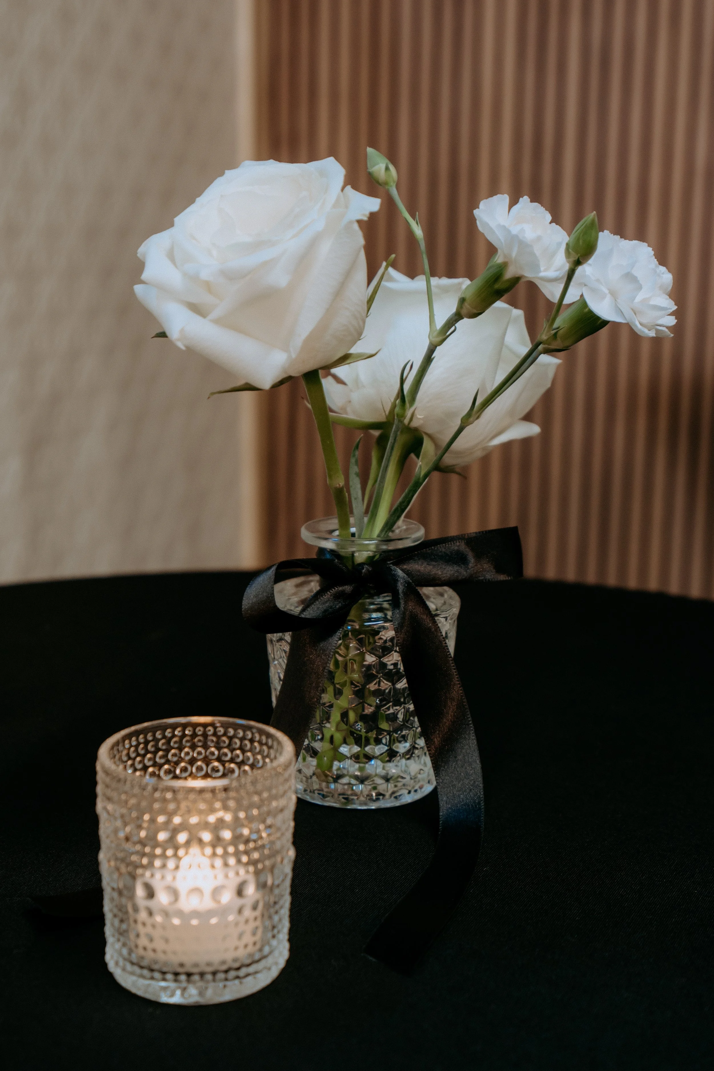 A small bouquet of white roses and carnations in a textured glass vase with a black ribbon tied around it, placed on a black table, with a candle holder in the foreground.