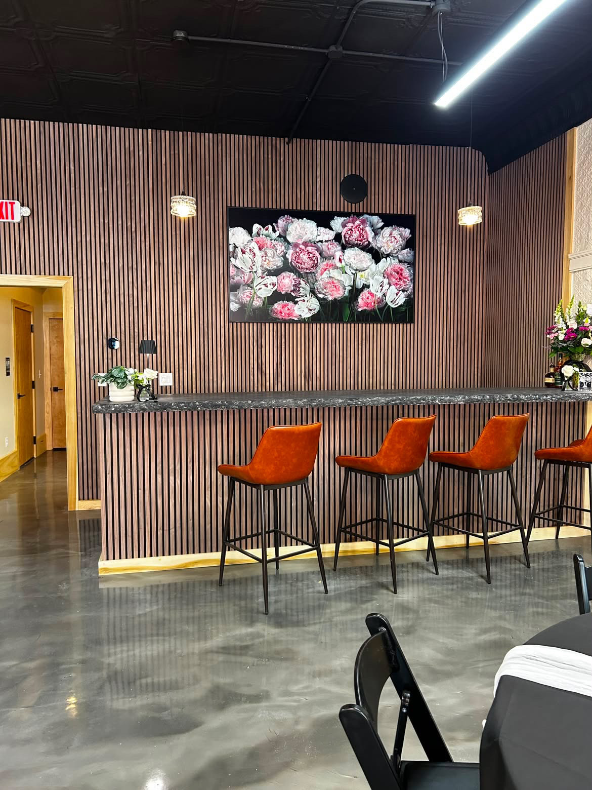 Interior of a modern bar with a wooden slat wall, a black marble countertop, and bar stools. A TV hangs on the wall above the bar, and there are decorative plants and flowers on the countertop.