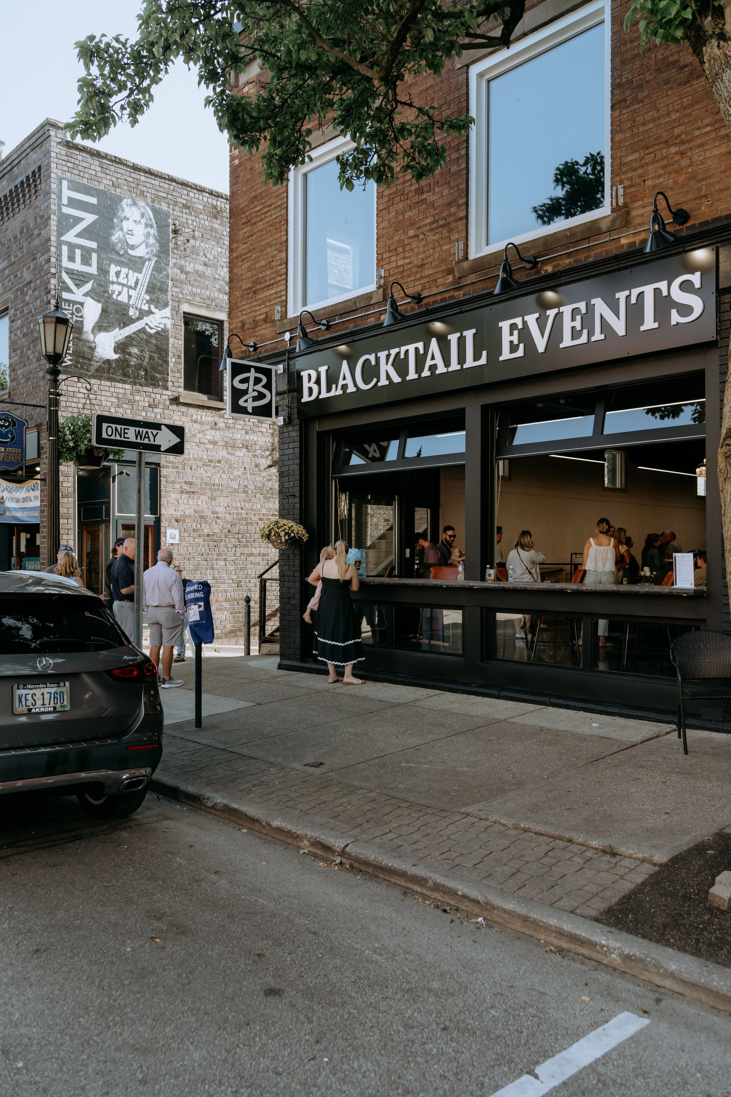Street scene with a brick building housing a venue called 'Blacktail Events', outdoor seating area with people, a brick wall with a black and white poster of a woman with a guitar, and a street sign pointing right with the words 'One Way'.