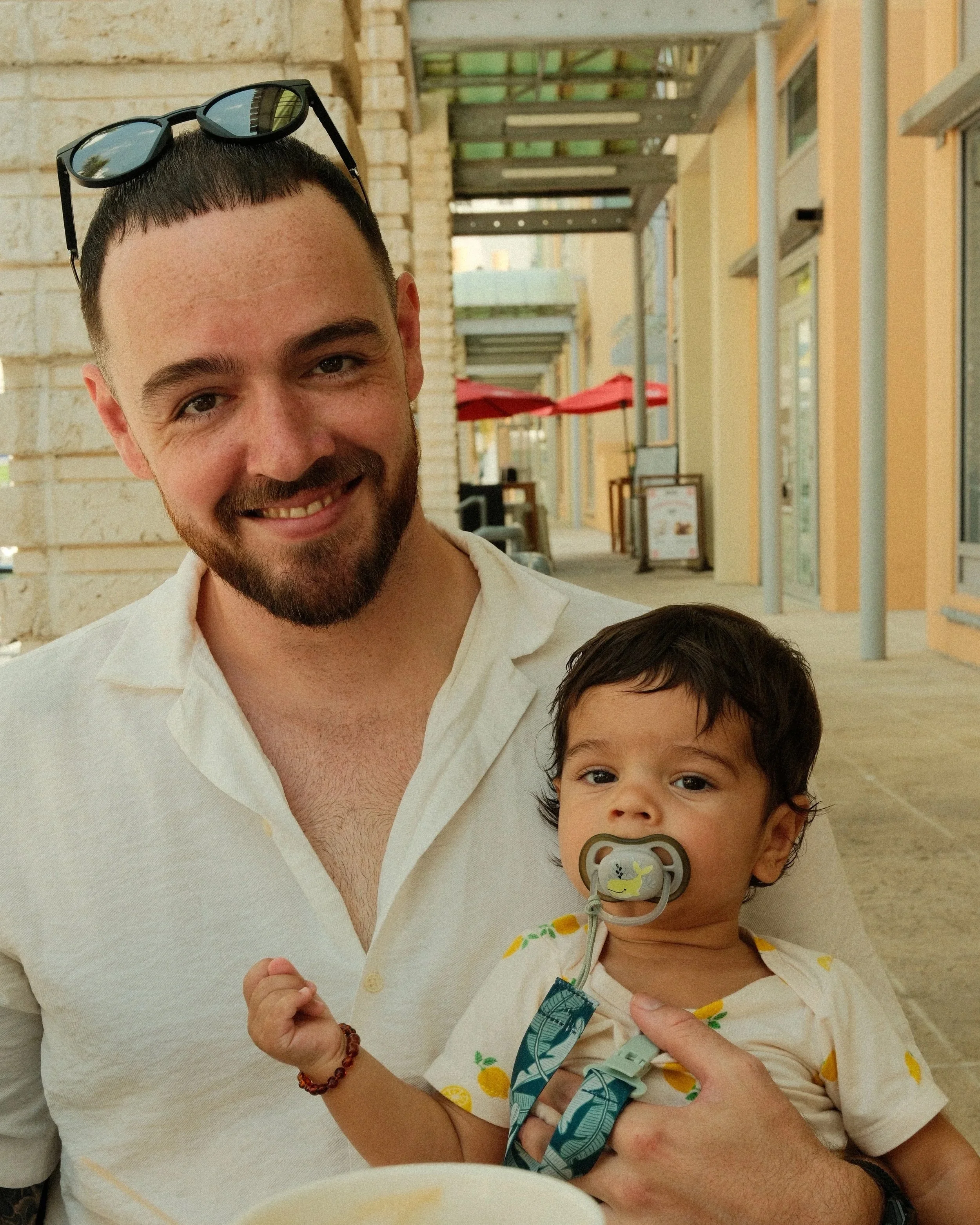 A smiling man with dark hair, beard, and sunglasses on his head holding a young child with a pacifier in his mouth, against a backdrop of outdoor shopping area with storefronts and red umbrellas.