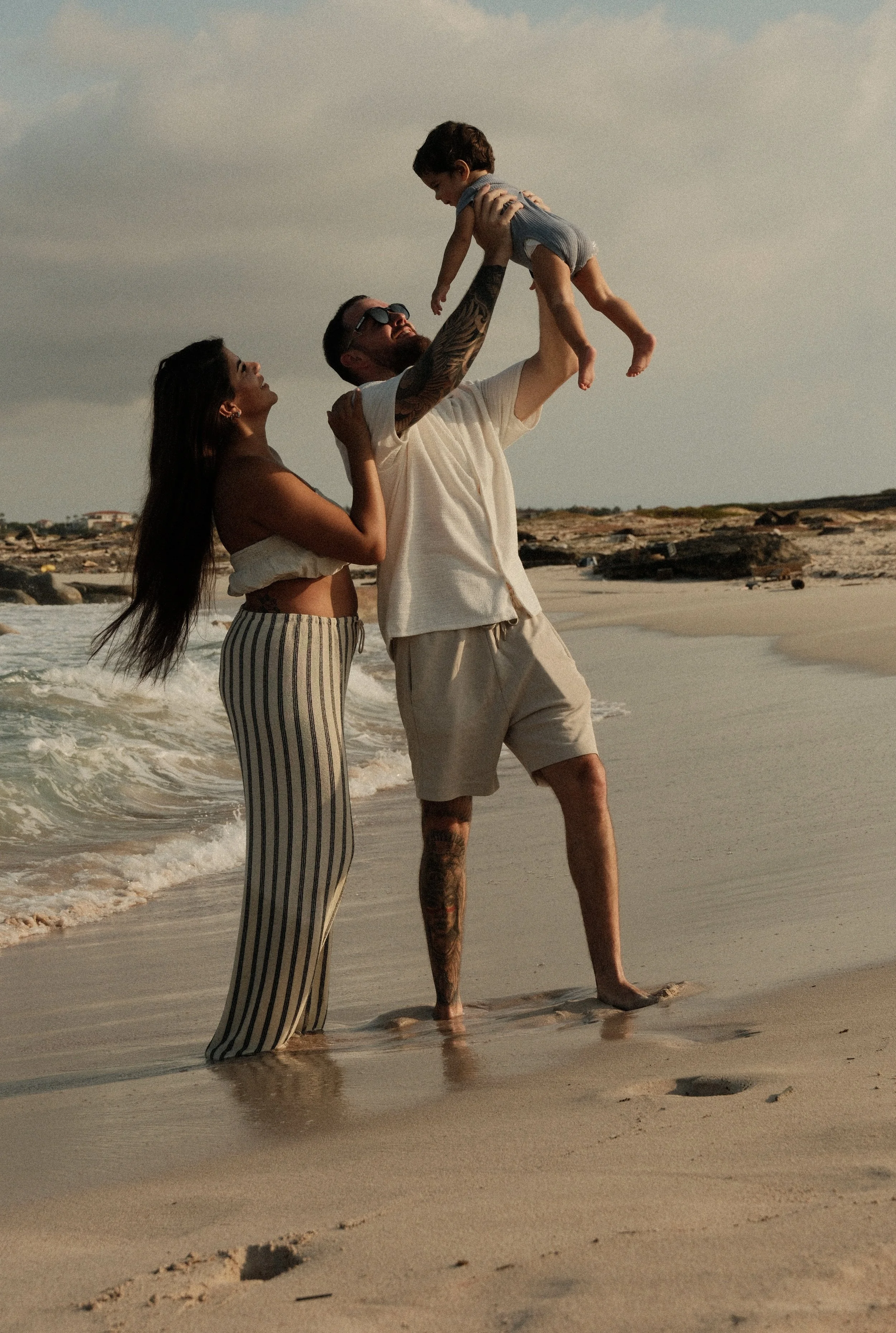 A family of three on the beach, with the father lifting a young child in the air, mother standing beside them, all enjoying a moment by the ocean with waves and a sandy shoreline.
