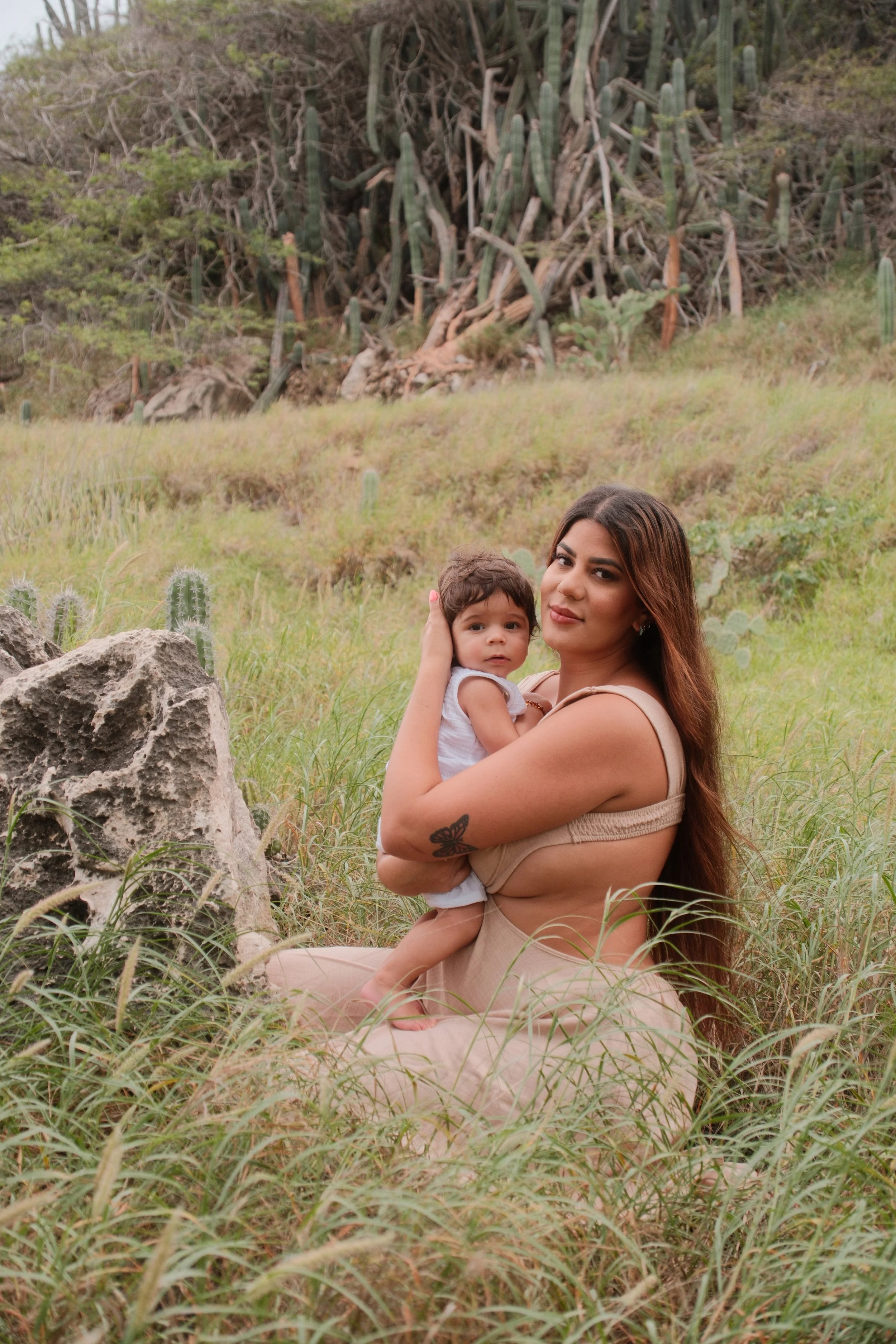 A woman sitting on the grass in a natural outdoor setting, holding a young child. The woman has long hair, a butterfly tattoo on her arm, and is wearing a beige dress. The child has short hair and is wearing a sleeveless top. There are rocks and cact