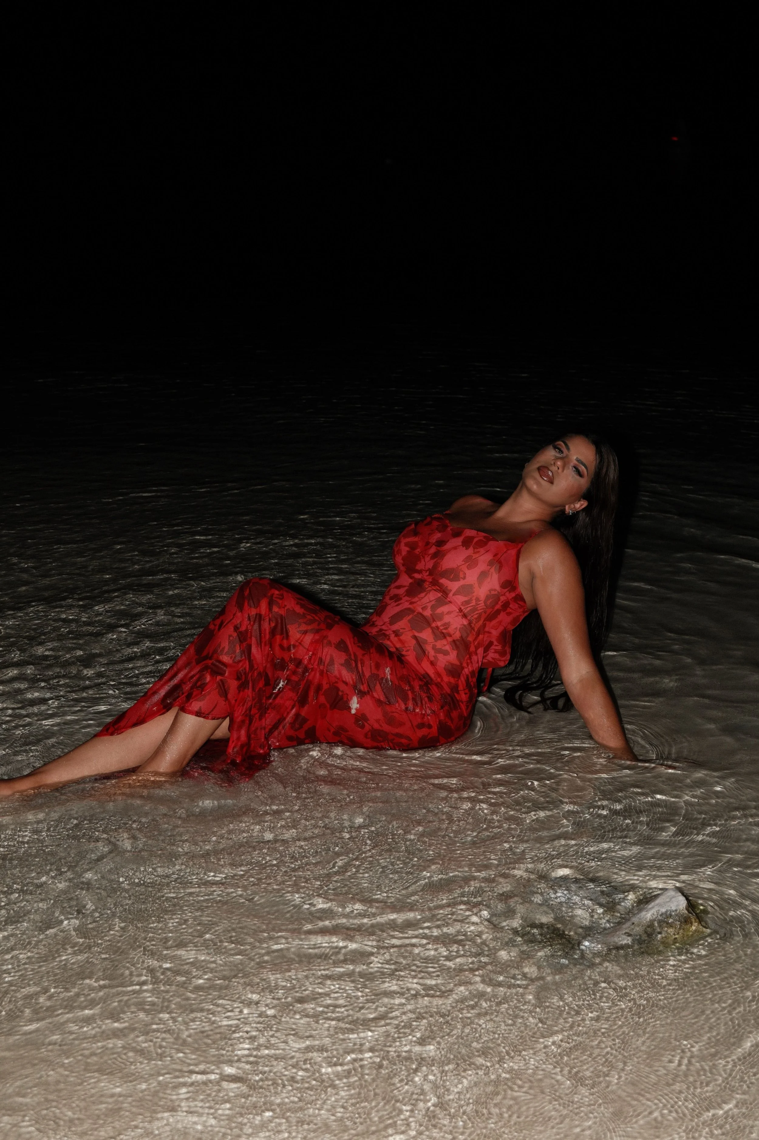 Woman in a red dress sitting in shallow water at night, with a dark background.