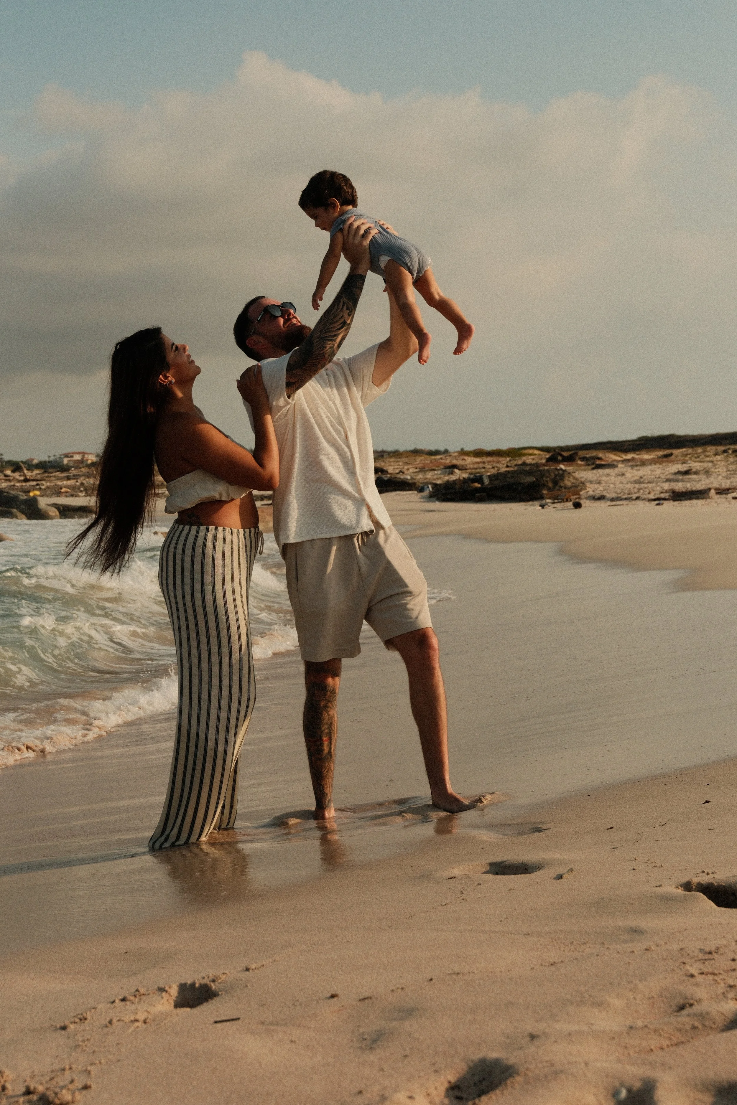 Family at the beach with a man lifting a child in the air, woman standing nearby, sandy shore, ocean waves, cloudy sky.