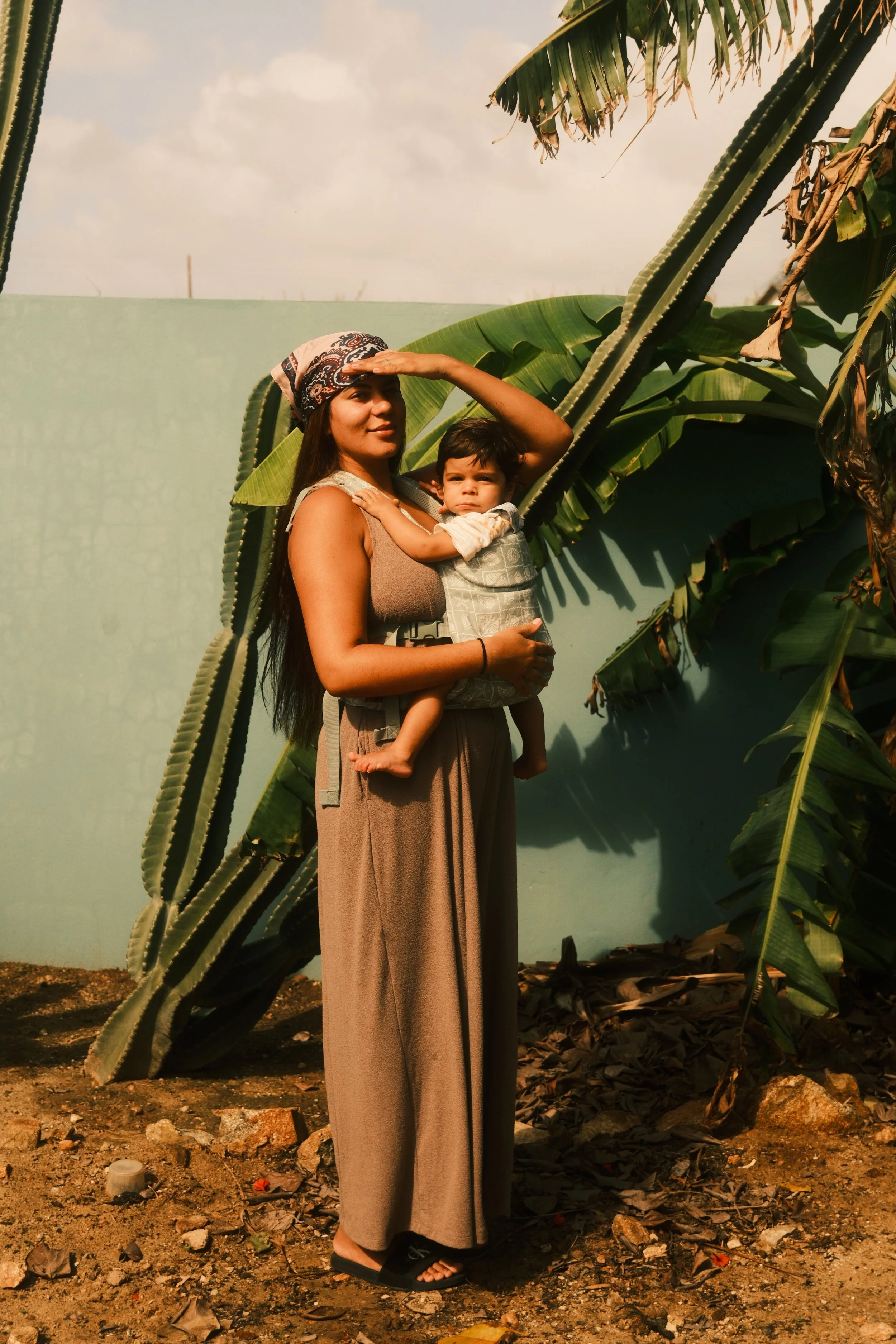 Woman carrying a young child in front of large banana plant leaves with a light blue wall in the background.