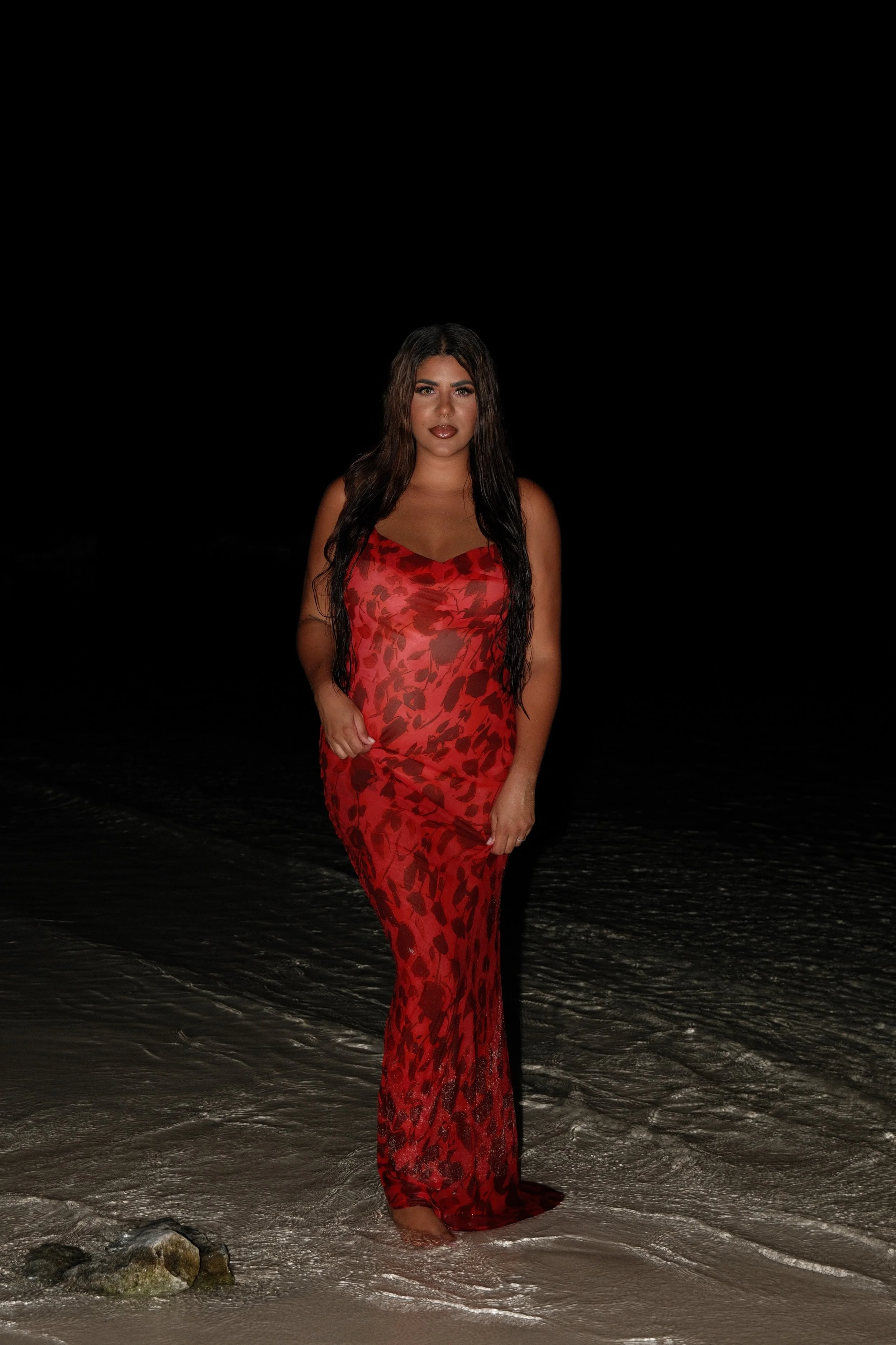 A woman standing on the beach at night, wearing a red and black patterned dress, with dark hair and makeup.