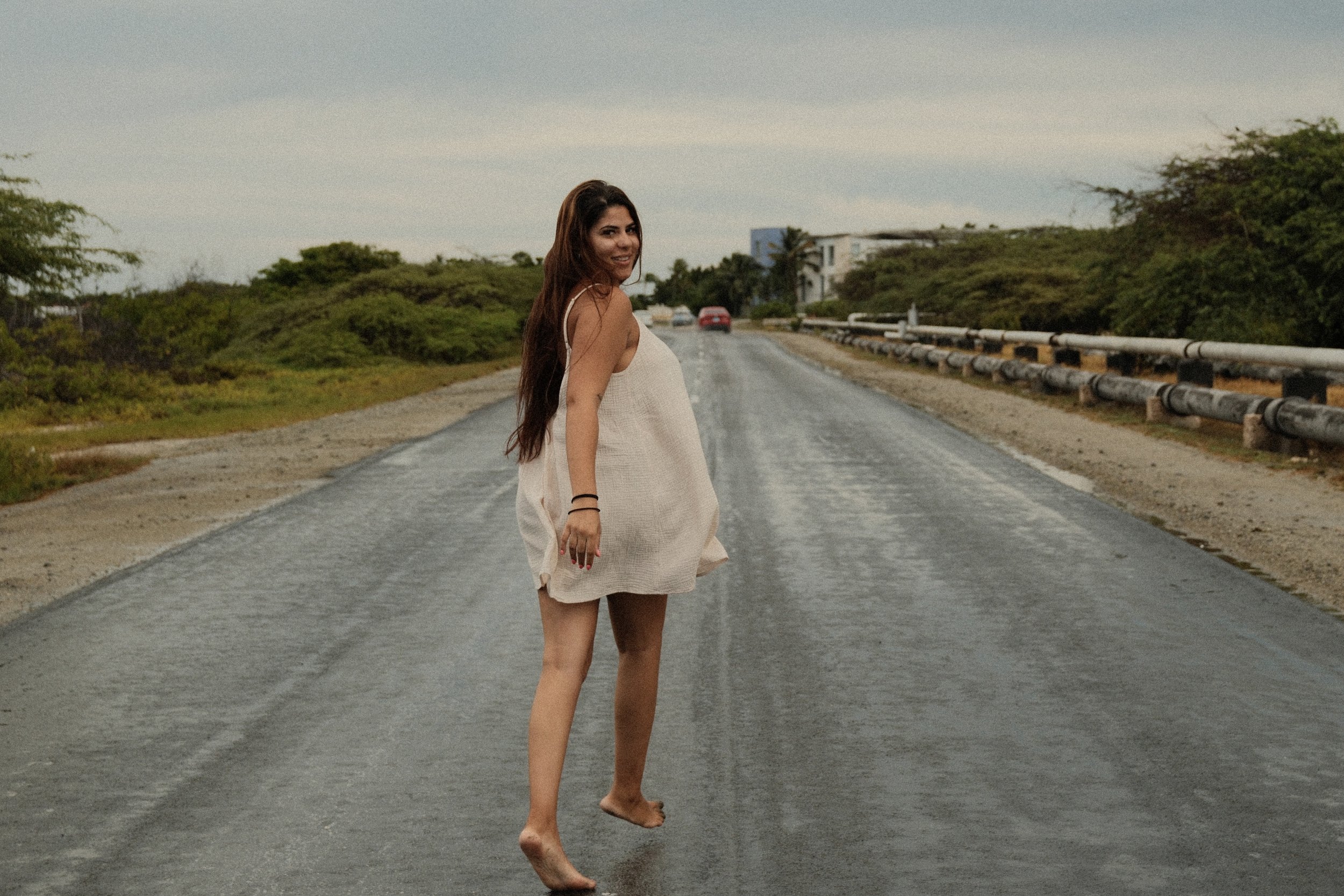 A woman in a white dress walking barefoot on a paved road with greenery on both sides and a cloudy sky overhead.