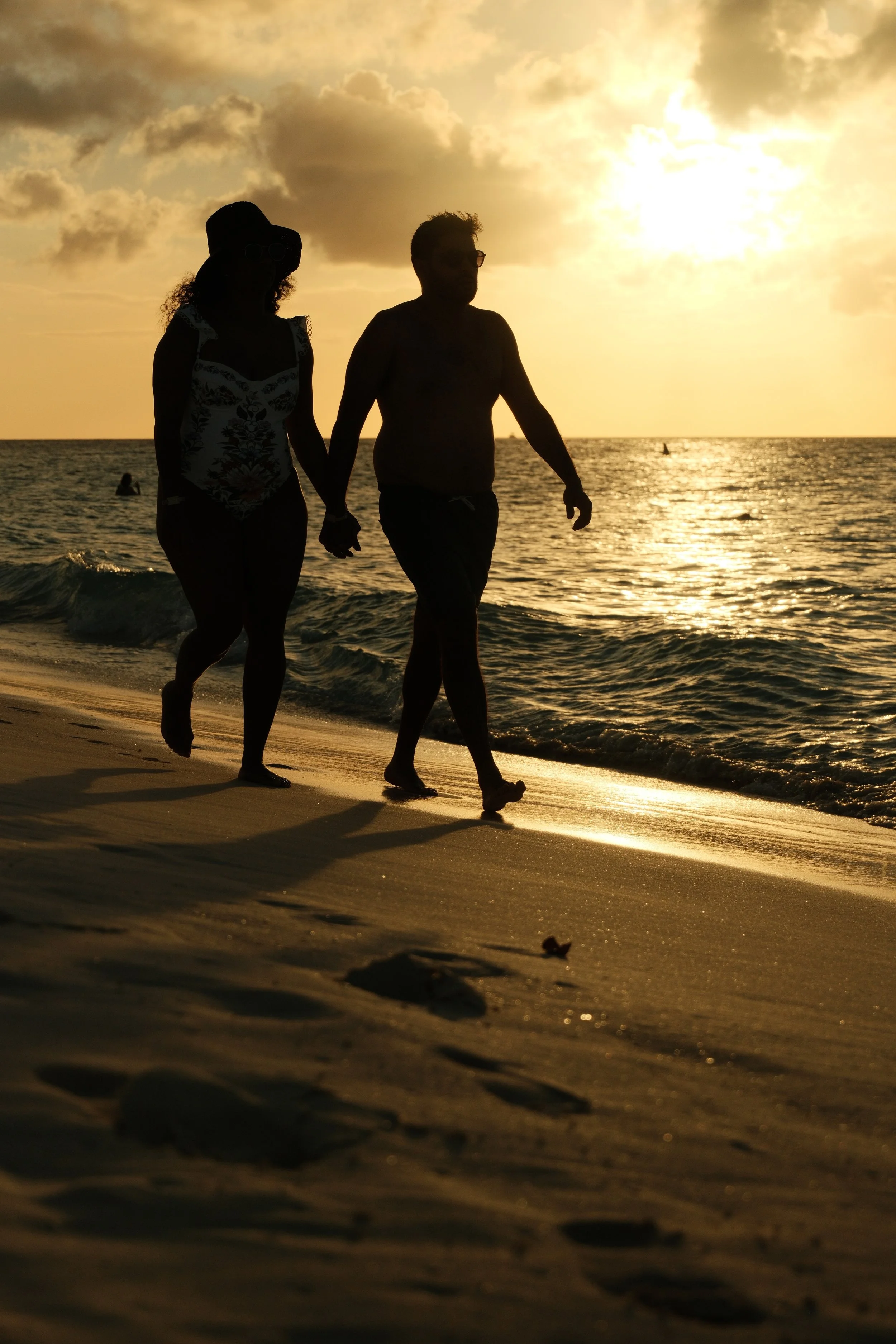 A silhouette of a man and woman holding hands walking along the beach at sunset, with the ocean waves and sky in the background.