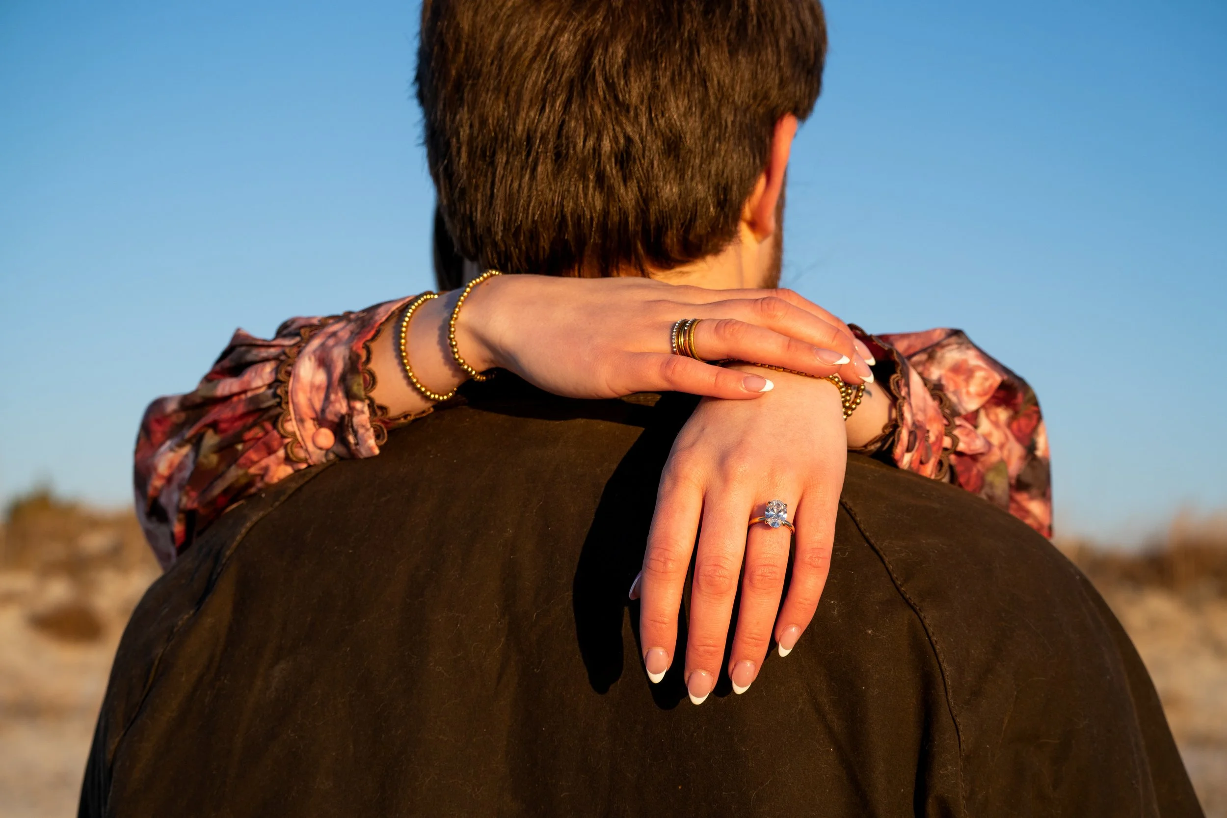 A woman with manicured nails and gold jewelry hugs a man from behind, focusing on her hands and their jewelry, with a clear blue sky in the background.